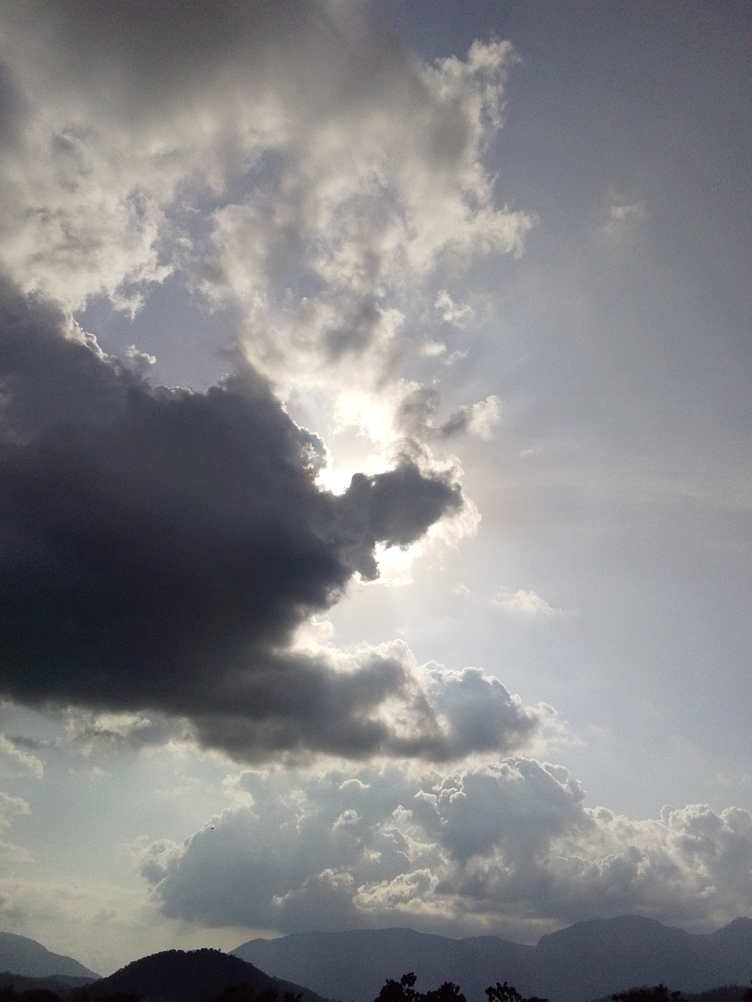 The image shows a dramatic sky with heavy, dark clouds dominating the left side, partially obscuring a bright sun. Lighter, more diffused clouds are visible throughout the frame, particularly towards the bottom. The sun's rays are breaking through in places, creating a spotlight effect. In the distance, silhouetted hills or mountains form the horizon line, with dark foliage visible in the foreground, suggesting a natural, possibly rural, setting. The overall impression is of a dynamic and possibly approaching storm, or the aftermath of one, with a contrast between light and shadow. The scene is a landscape view. There are no people, specific objects, or actions depicted. The location is identified as Serti, Nigeria.