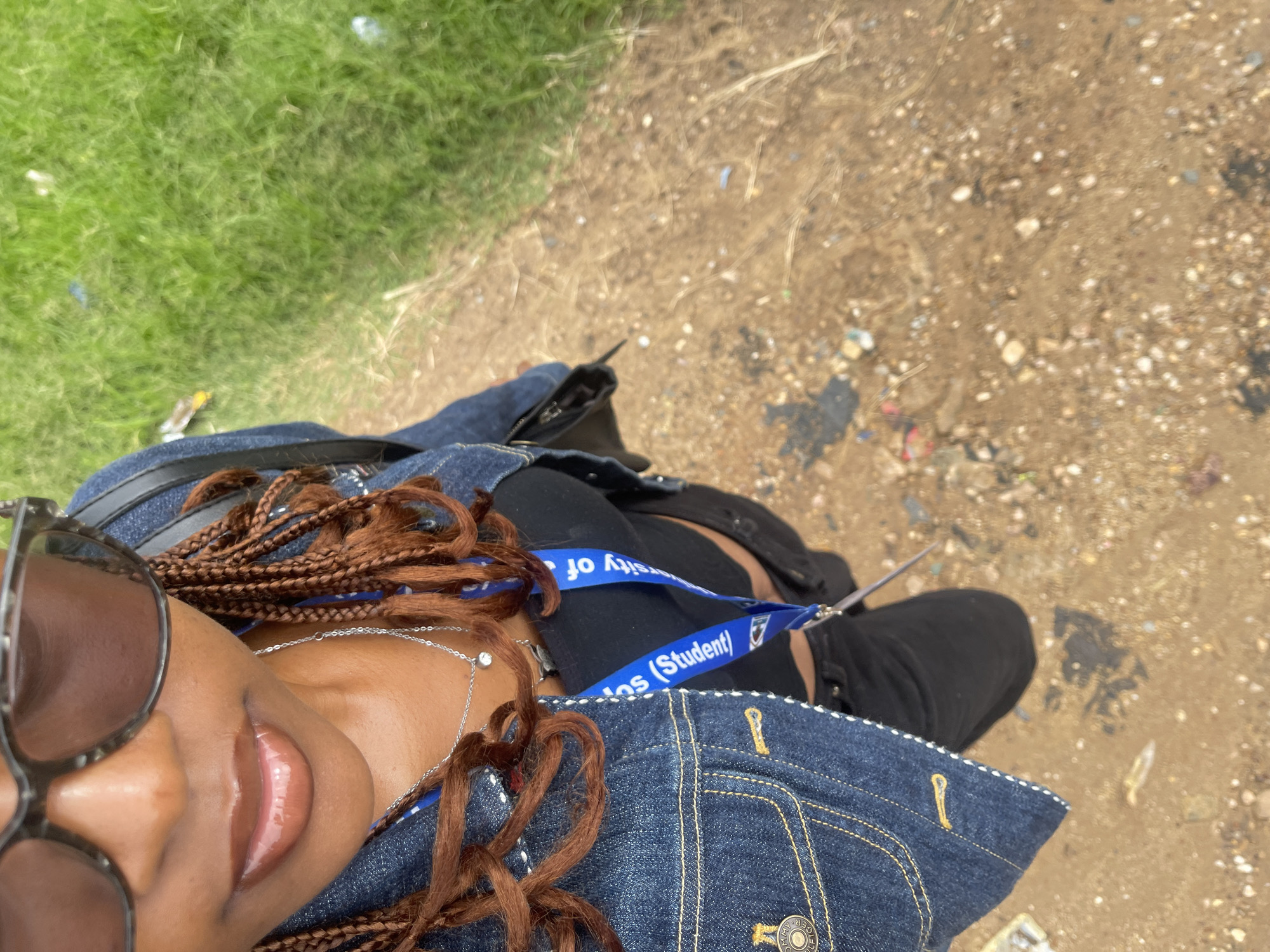 A woman is captured in a high-angle, close-up shot, from the chest up. She is wearing large, dark sunglasses, a denim jacket over a black top, and black pants. A blue lanyard with white text, appearing to be for a university student, hangs around her neck. Her hair is styled in brown braids that fall forward. She is accessorized with a silver necklace featuring a small pendant.
The background consists of a dry, dirt path scattered with small stones and debris, bordering a strip of vibrant green grass. The scene appears to be outdoors, likely in a semi-urban or rural setting. The specific location is identified as Rafin Par, Nigeria. The lighting suggests it is daytime, with the sun providing natural illumination. The overall composition is casual and personal, possibly a selfie.