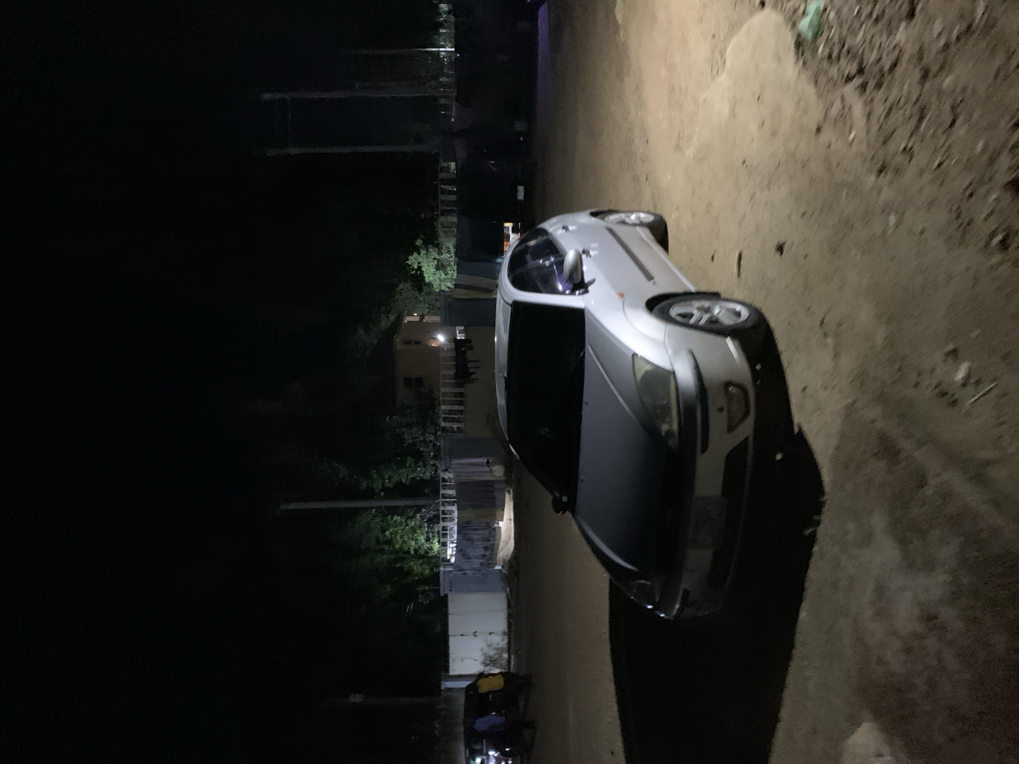 The image depicts a silver car parked on a dirt or gravel road at night. The location appears to be in or near a residential area, possibly in the city of New Stadium, Nigeria, given the surrounding buildings and infrastructure. The lighting is dim, with the car and the immediate area illuminated by what seems to be overhead or street lighting. The rest of the scene is shrouded in darkness. No people are distinctly visible.