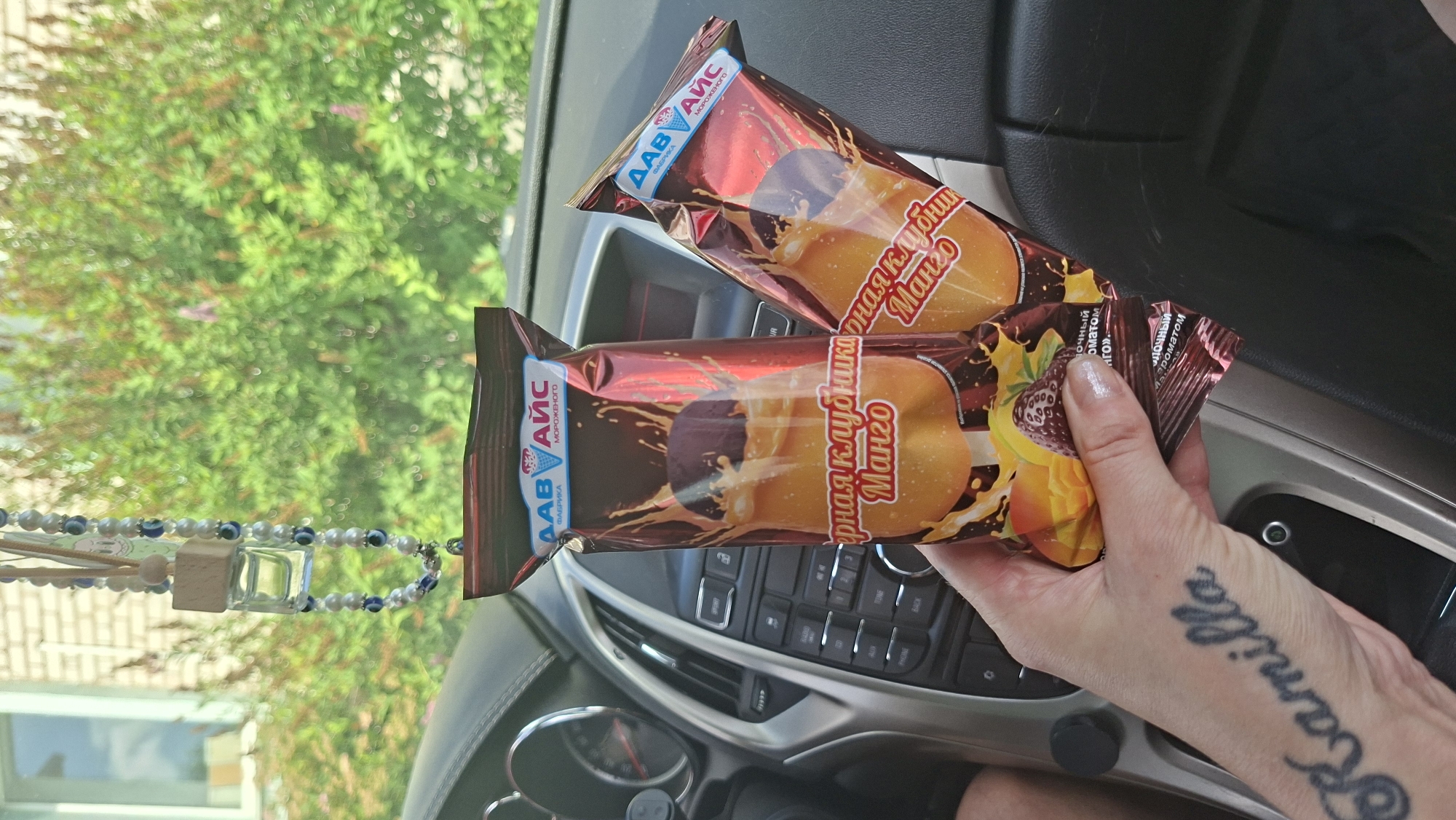 In Myadzyel, Belarus, a hand with the tattooed name "Kamilla" on the wrist holds two ice cream bars in colorful packaging. The bars are depicted with images of dripping chocolate, splashing liquid, and fruit, suggesting flavors like strawberry and mango. They are positioned in the foreground, partially obscuring the dashboard of a car. The car's interior features a dark dashboard with visible buttons and dials. Through the car's windshield, lush green foliage and a portion of a building are visible, indicating an outdoor setting. A beaded car air freshener with a wooden top and blue and white beads hangs from the rearview mirror, adding a decorative element to the scene. The overall impression is a candid snapshot of a person holding ice cream in a car on a sunny day.