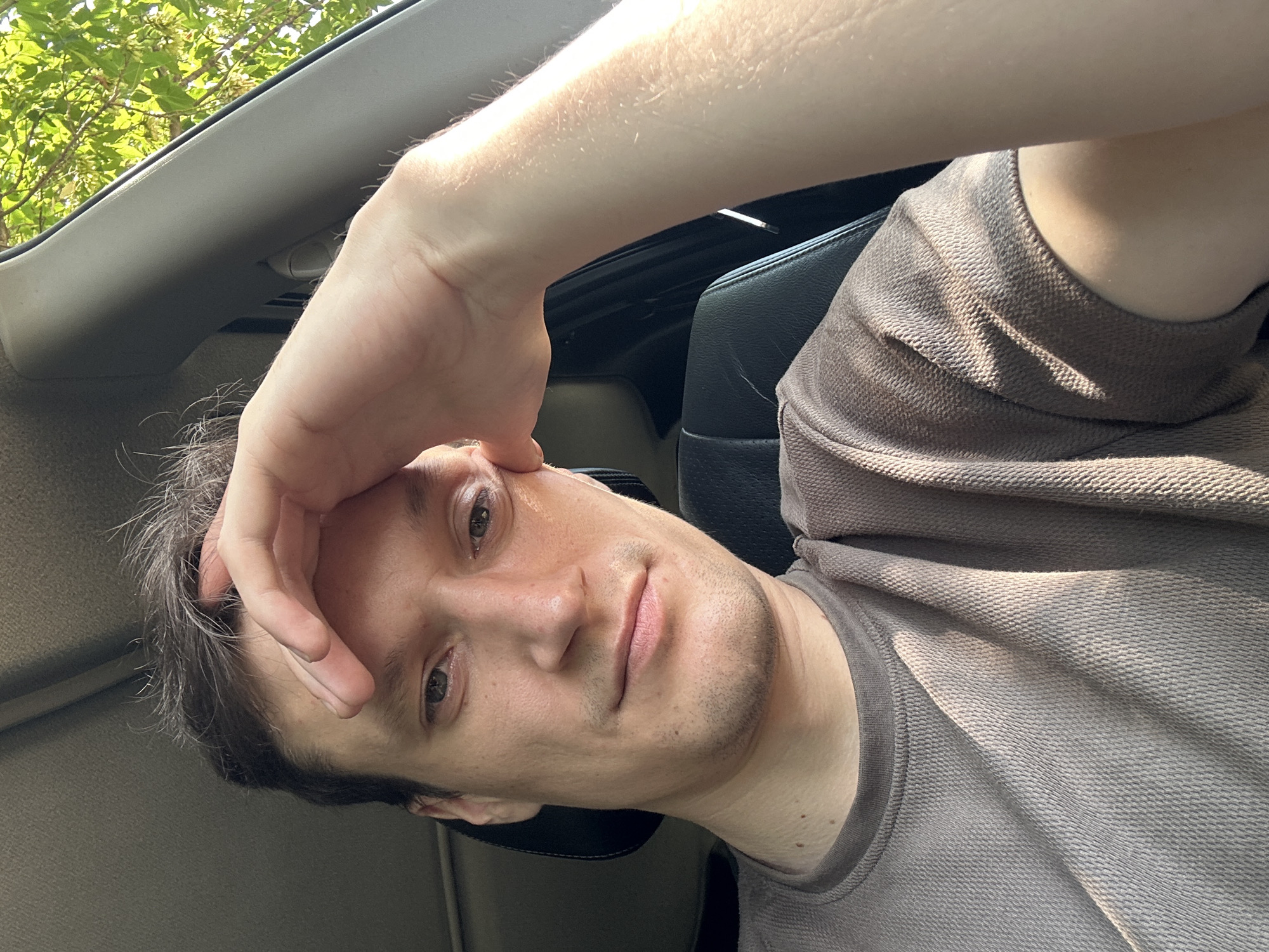 A young man is in the driver's seat of a car. He is looking at the camera and holding his forehead with his right hand. His hair is dark brown and messy. He is wearing a brown t-shirt. The car interior is visible behind him, including a black leather seat and a beige headliner. Sunlight is streaming in from the right side of the car, illuminating his arm and face. Green leaves from trees are visible outside the car window. The man's expression appears to be one of fatigue or contemplation.