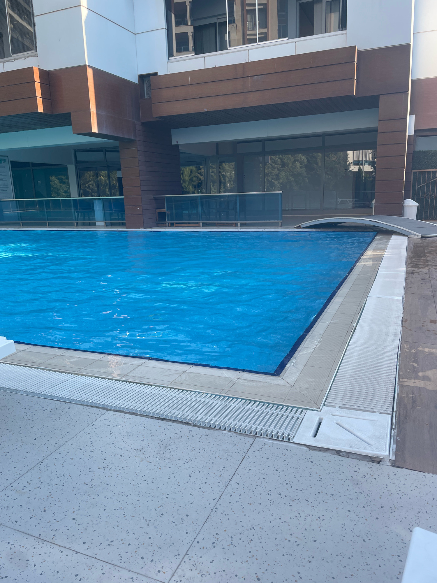The image shows a swimming pool covered with a bright blue tarp. The pool is located outdoors, adjacent to a modern building with a facade of white panels and brown wooden accents. There are large windows on the building, reflecting trees and other structures. The pool area is surrounded by light-colored tiled pavement and drainage grates. In the background, a part of a retractable pool cover is visible, arching over the water. The setting appears to be a sunny day. The specific location in Yunus Emre, Turkey, is not discernible from the image itself but is provided as context.