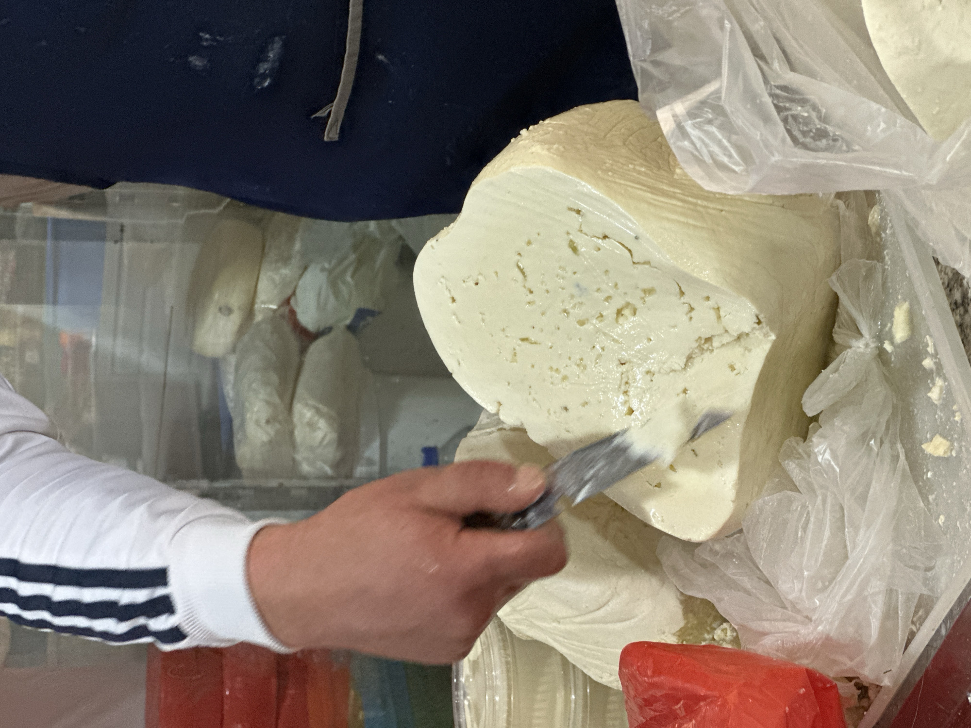 A person is cutting a large block of white cheese with a knife. The person is wearing a white long-sleeved shirt with black stripes on the sleeve. The cheese is sitting on top of a pile of other cheeses wrapped in clear plastic bags. The background appears to show a refrigerated display case filled with more cheeses. There are also red and blue objects in the lower left corner. The location is likely a cheese shop in the city of Yerevan, Armenia.