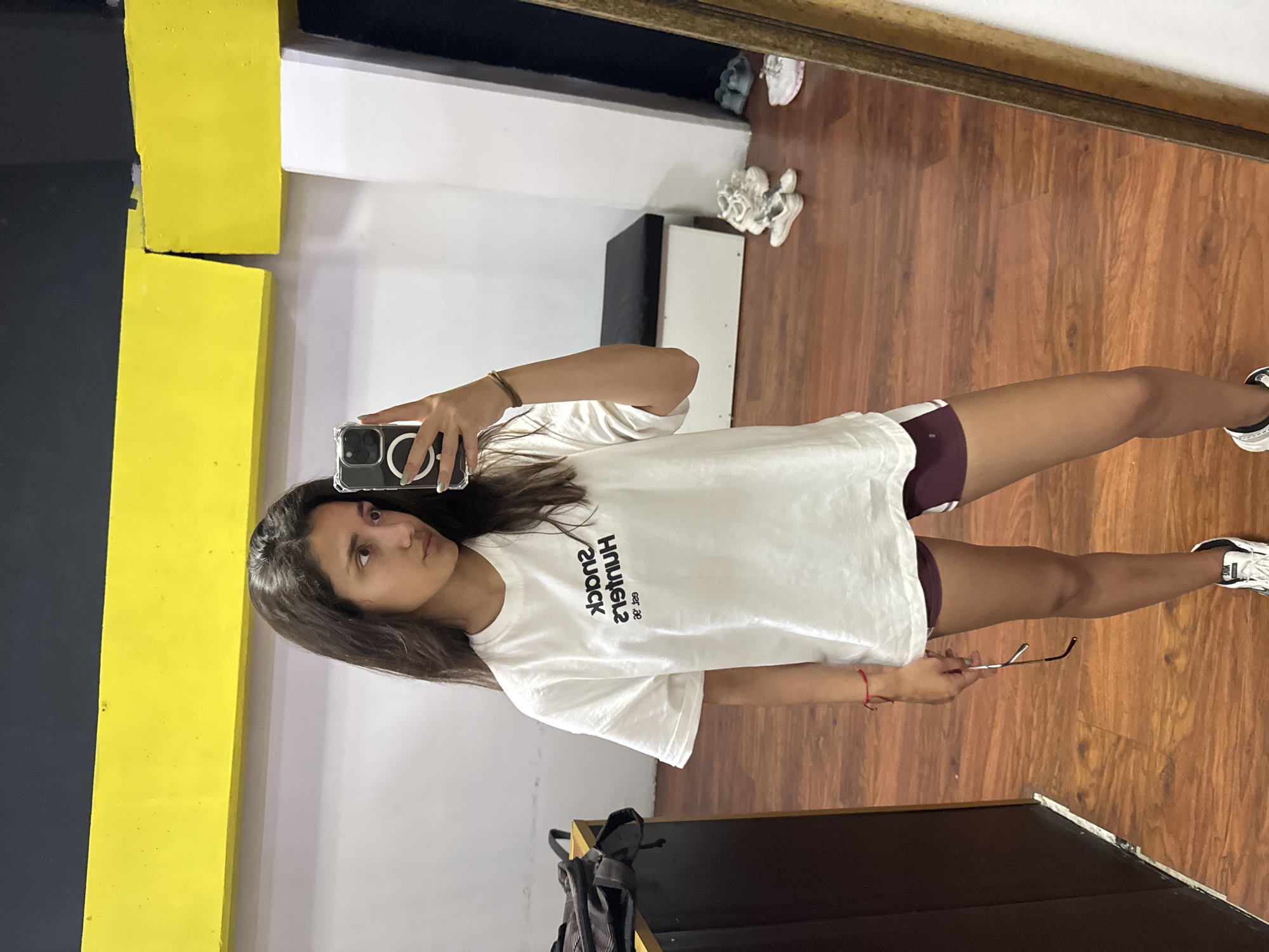 A young woman is taking a mirror selfie in what appears to be an indoor space. She is wearing a white t-shirt and maroon shorts. In her right hand, she holds a pair of glasses, while her left hand holds a smartphone. The environment shows a wooden floor and painted walls with black, yellow, and white sections. Several pairs of shoes are visible near a white cabinet or furniture piece. The presence of a mirror indicates she could be in a changing room, gym, or studio. There is no information to confirm that the image was taken in the city of Bar, Montenegro.