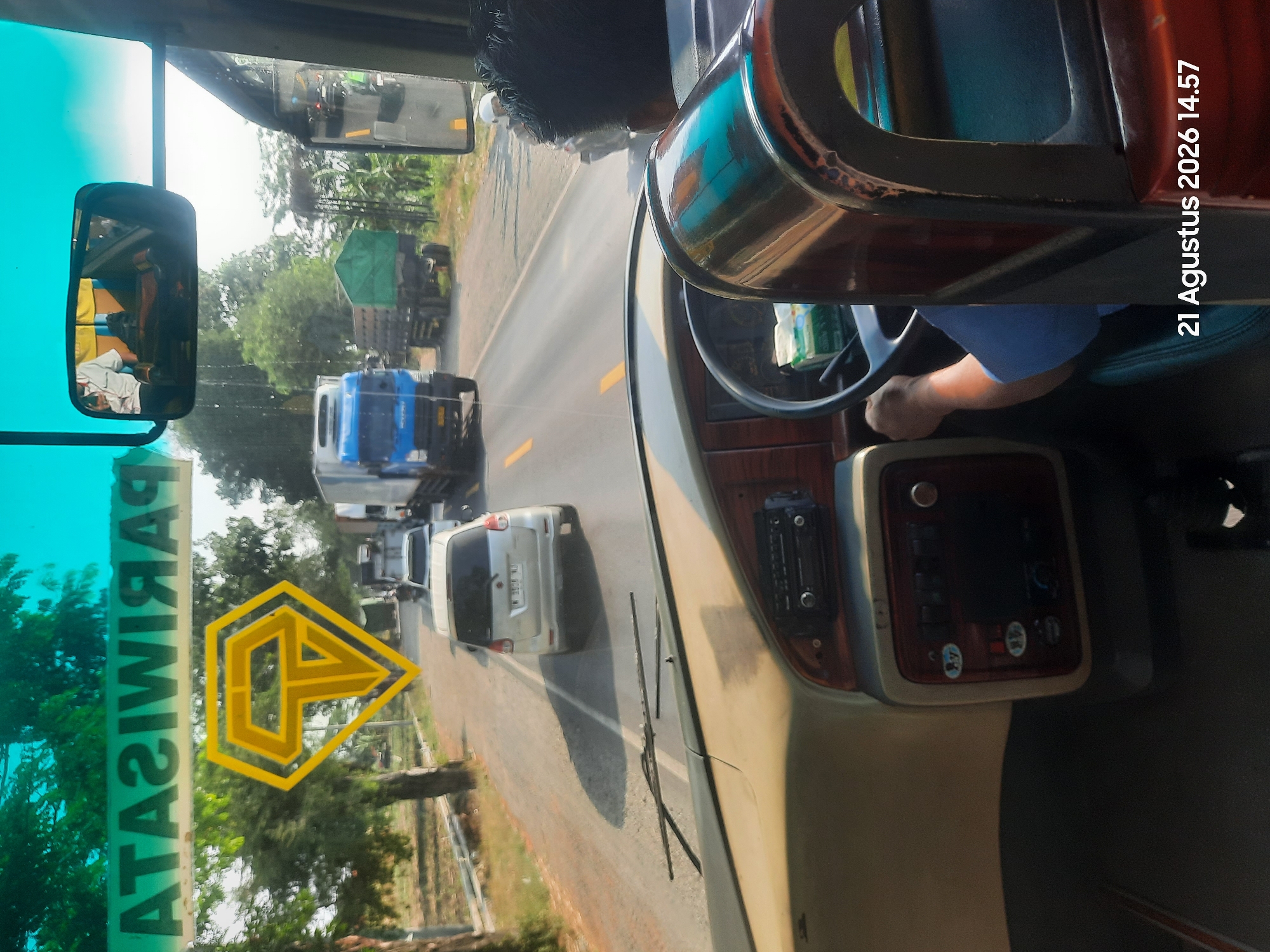 The image, captured on August 21, 2026, at 14:57, provides an interior view from the driver's seat of a bus traveling on a road in Sukokerto, Indonesia. The windshield shows a bright, sunny day with a blue sky and green trees. A yellow, diamond-shaped logo with a stylized 'T' is visible on the windshield. Outside, the road is lined with trees and there are several vehicles including a white car, a blue truck, and a green truck. The bus interior includes a dashboard with a radio and other controls, and a steering wheel. The driver's hands are visible on the steering wheel, and a person wearing a blue shirt is seated in the driver's seat. A side mirror reflects a person in a white shirt. The overall scene depicts a journey on a rural or suburban road in Indonesia.