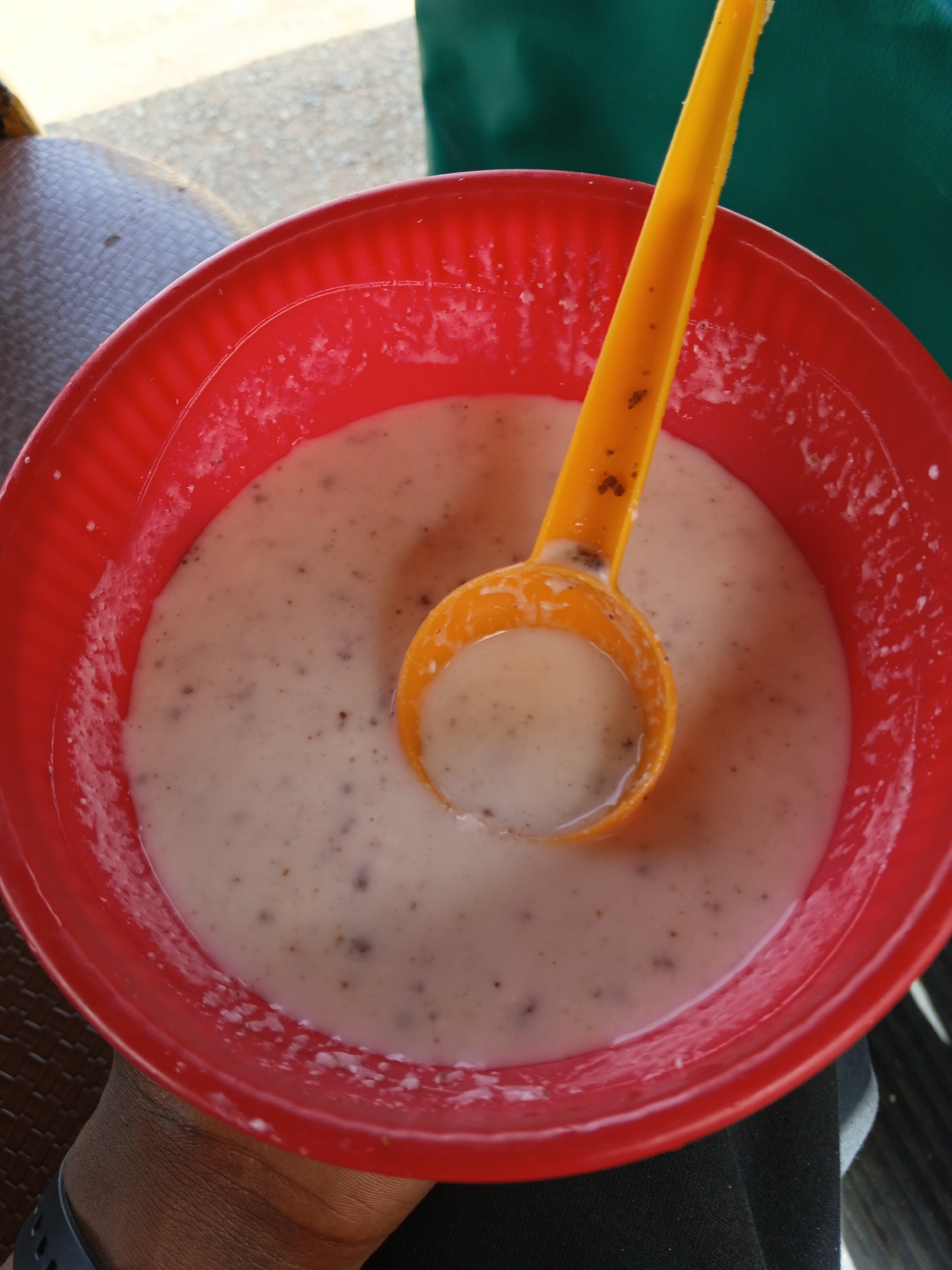 This image is a close-up, overhead shot of a red plastic bowl filled with a creamy, light-colored substance speckled with dark flecks. An orange plastic spoon is partially submerged in the mixture, with its bowl scoop facing upwards. The spoon appears to have some of the food clinging to it. The red bowl has a ribbed texture around its rim. The background is blurred, showing what appears to be a textured brown surface on the left and a dark green fabric on the right. A person's hand, with dark skin and a watch on the wrist, is visible in the lower left corner, holding the bowl. The lighting suggests it could be daytime, possibly outdoors or in a well-lit indoor space. No text is visible in the image.