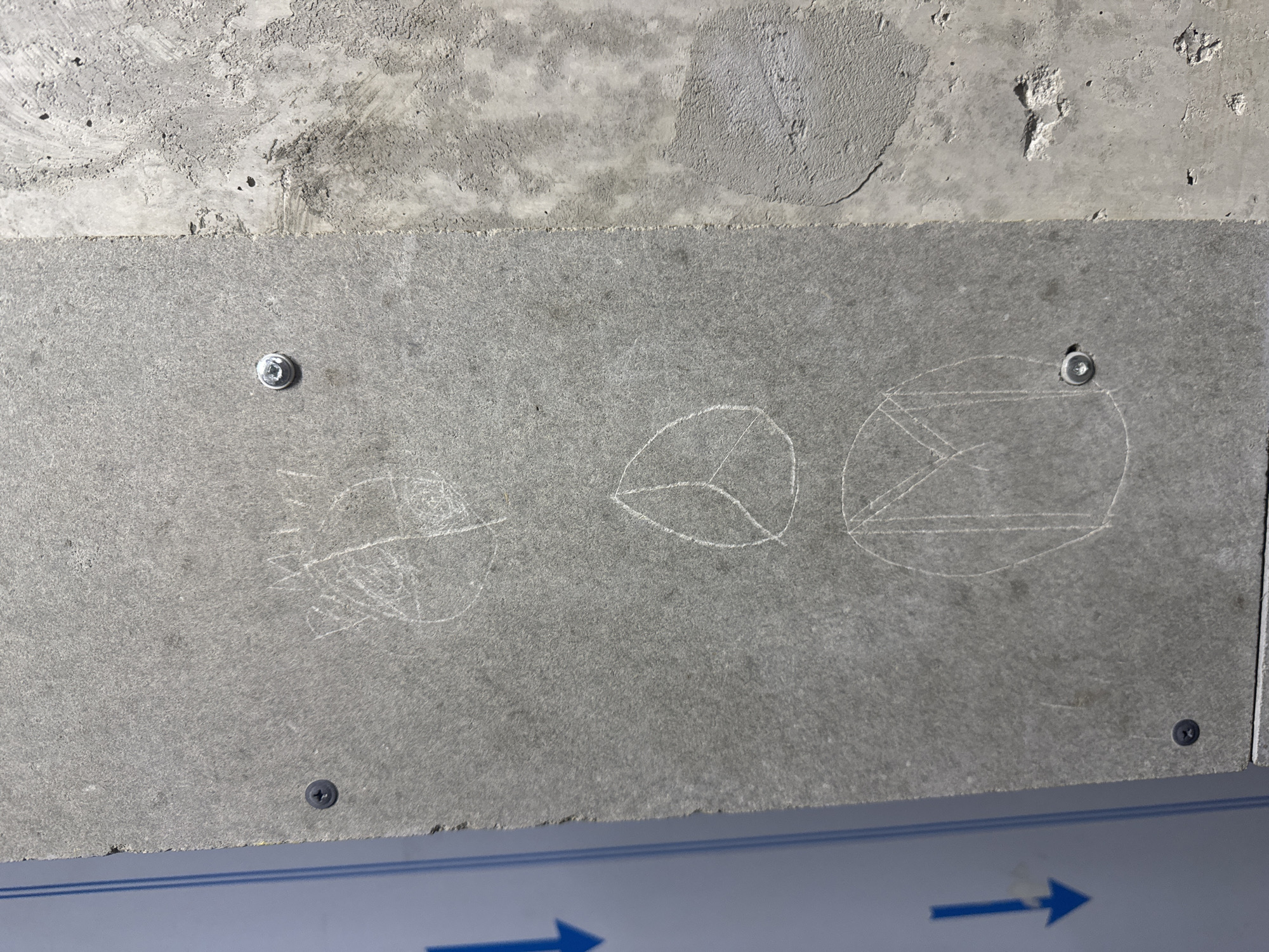 The image depicts a gray rectangular panel affixed to a rough concrete ceiling with screws. Three crude drawings are etched onto the panel using a white material. The first drawing resembles a face with radiating lines. The second drawing appears to be two intersecting teardrop shapes. The third drawing is a circle enclosing a triangle and a rectangle. Below the panel is a section of a metallic blue structure with white arrows. The location in the city of Unknown, Unknown is not apparent from the image.