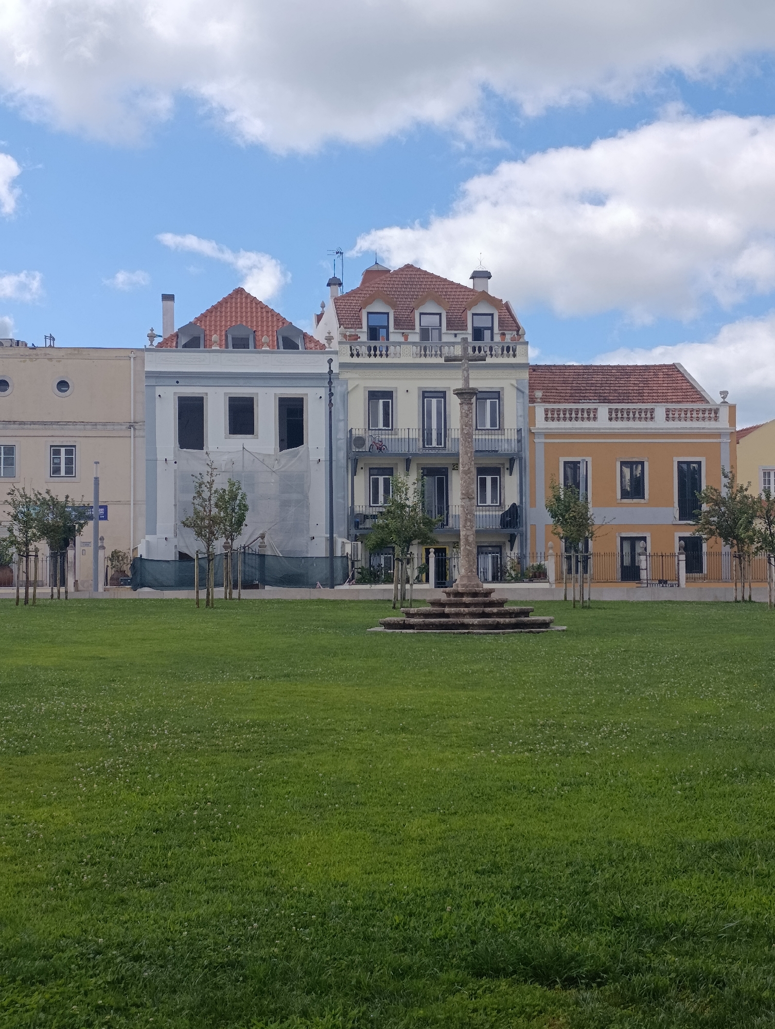 The image depicts a sunny day in Setúbal, Portugal, at Praça de Bocage. In the foreground, a vast expanse of well-maintained green lawn fills the lower half of the frame, dotted with small white wildflowers. The middle ground features a historic stone monument, likely a cross, with a multi-tiered base, standing in the center of a circular paved area. Behind the monument, a row of colorful buildings lines the square. From left to right, the buildings include a pale yellow structure with circular windows, a light blue building undergoing renovation with white sheeting covering its facade, a yellow building with blue accents and balconies, and a partially visible orange building. A line of slender trees with sparse foliage is planted in front of the buildings, separated by low metal fencing. The sky above is a bright blue, adorned with fluffy white clouds. No people are visible in the scene.