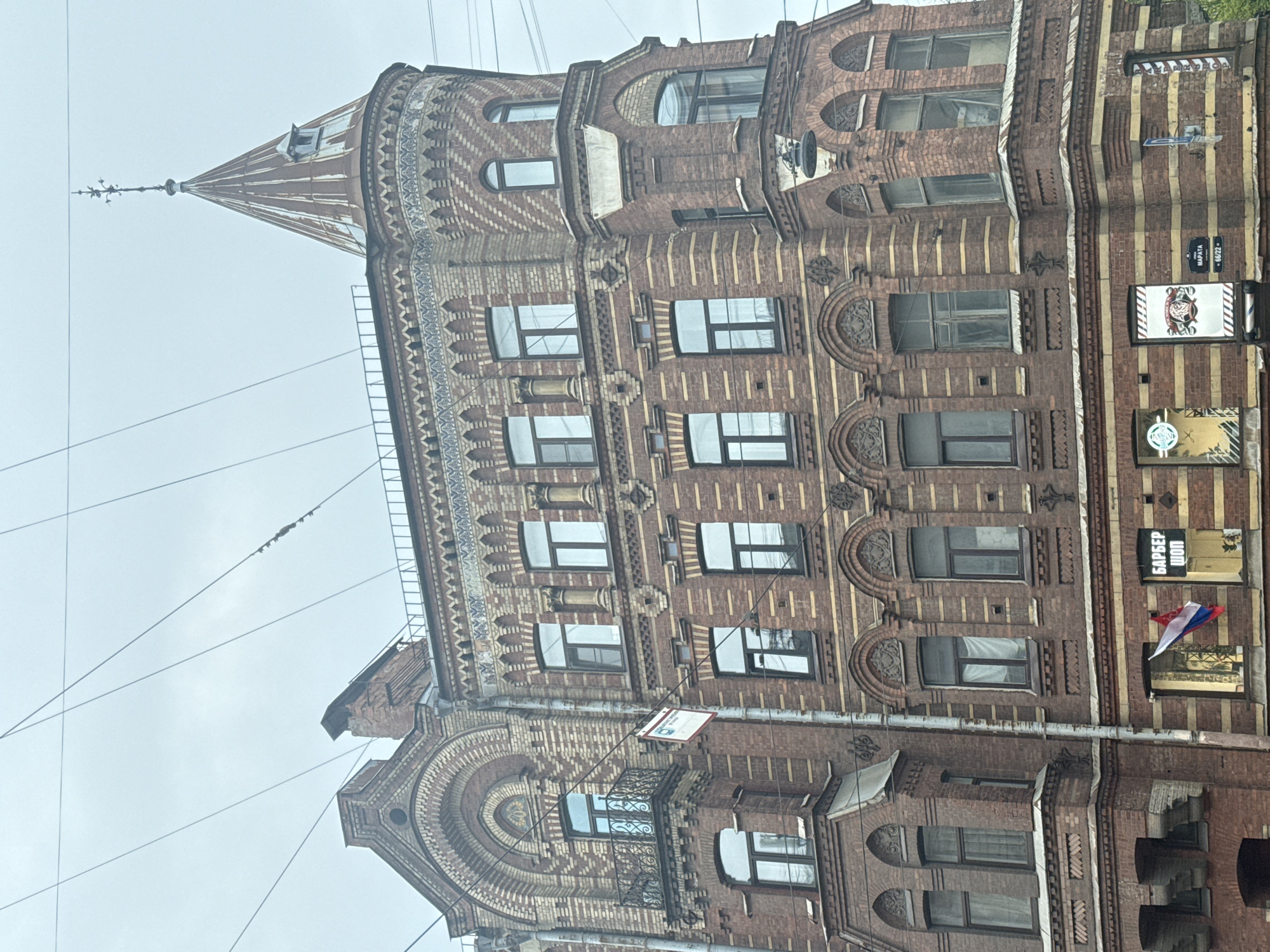 The image shows a multi-story brick building with intricate architectural details in Saint Petersburg, Russia. The building features various window shapes and sizes, decorative brickwork, and a conical tower on one corner. Electrical wires are visible against a cloudy sky. The building's facade also includes signage for businesses, including what appears to be a barbershop, and a small Russian flag.