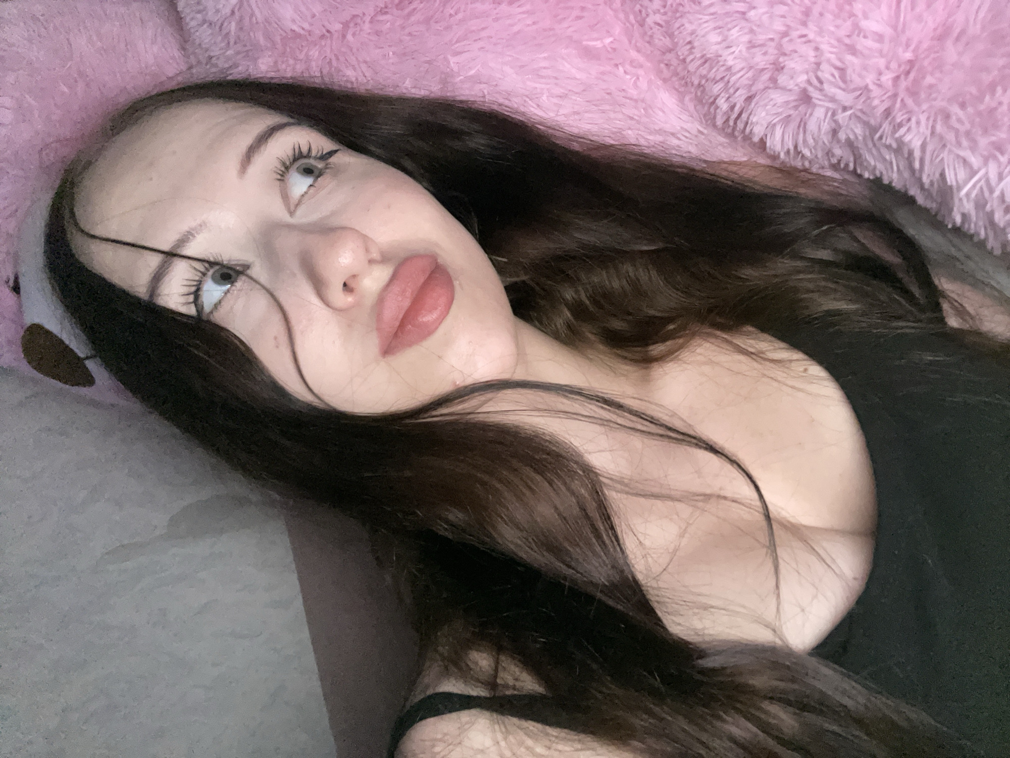 A young woman with long dark hair and large blue eyes is lying down, looking to her right. She has full lips and is wearing a black top. She is surrounded by a fluffy pink material, possibly a blanket or stuffed animal. The background appears to be a wall with some texture. The scene is dimly lit. There is no discernible information about the location in Votkinsk, Russia.