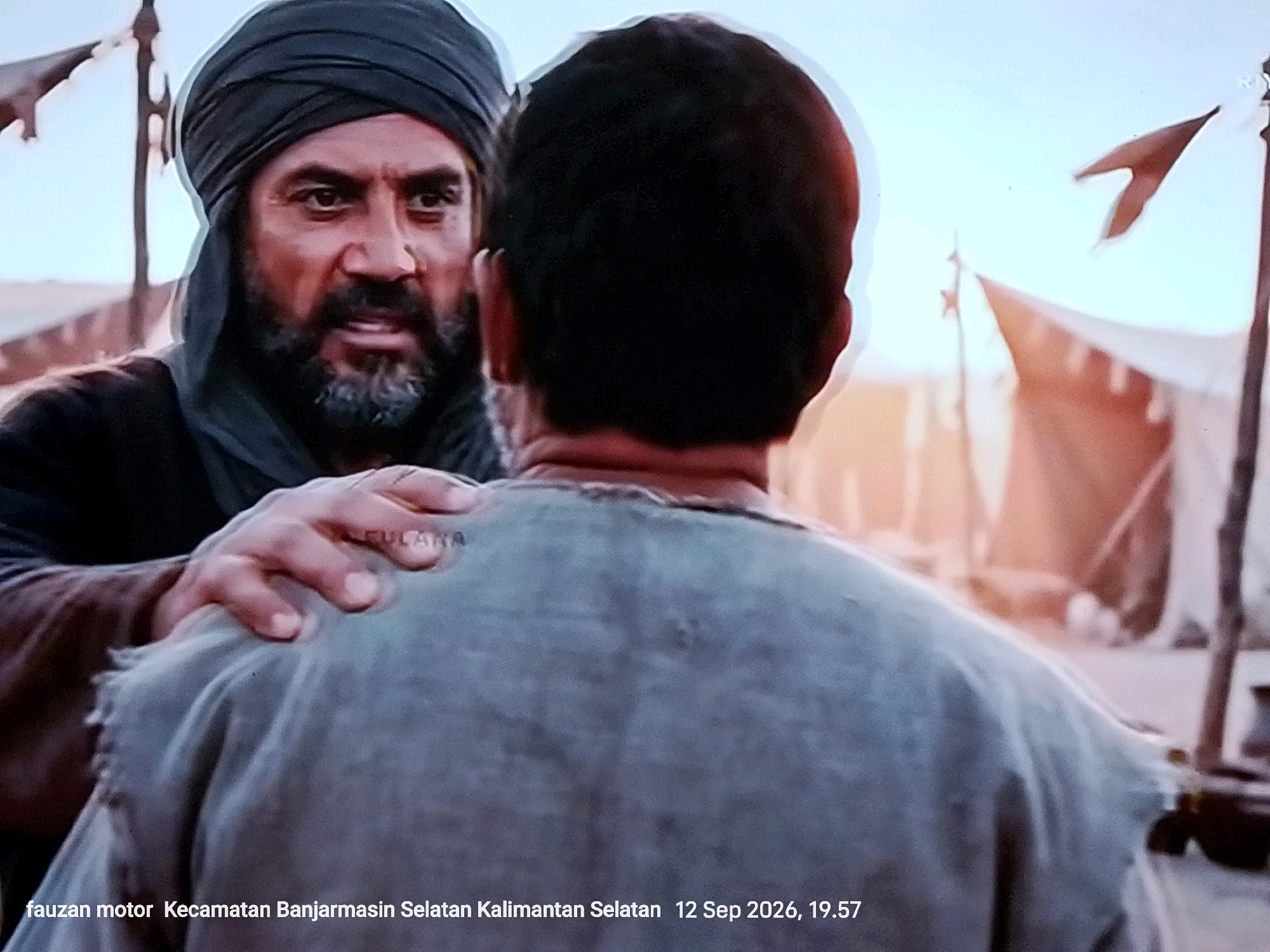 The image shows two men in what appears to be a desert encampment. One man, with a dark turban and beard, is looking intensely at the other man, whose back is to the camera. The man in the foreground has his hand on the shoulder of the man in the background, and there appears to be text on his hand, possibly a watermark. Tents and flags are visible in the background, suggesting a temporary settlement. The bottom of the image has text overlay, indicating "fauzan motor Kecamatan Banjarmasin Selatan Kalimantan Selatan 12 Sep 2026, 19.57". This suggests the location is in the South Banjarmasin District of South Kalimantan, Indonesia, and the image was captured on September 12, 2026, at 7:57 PM.