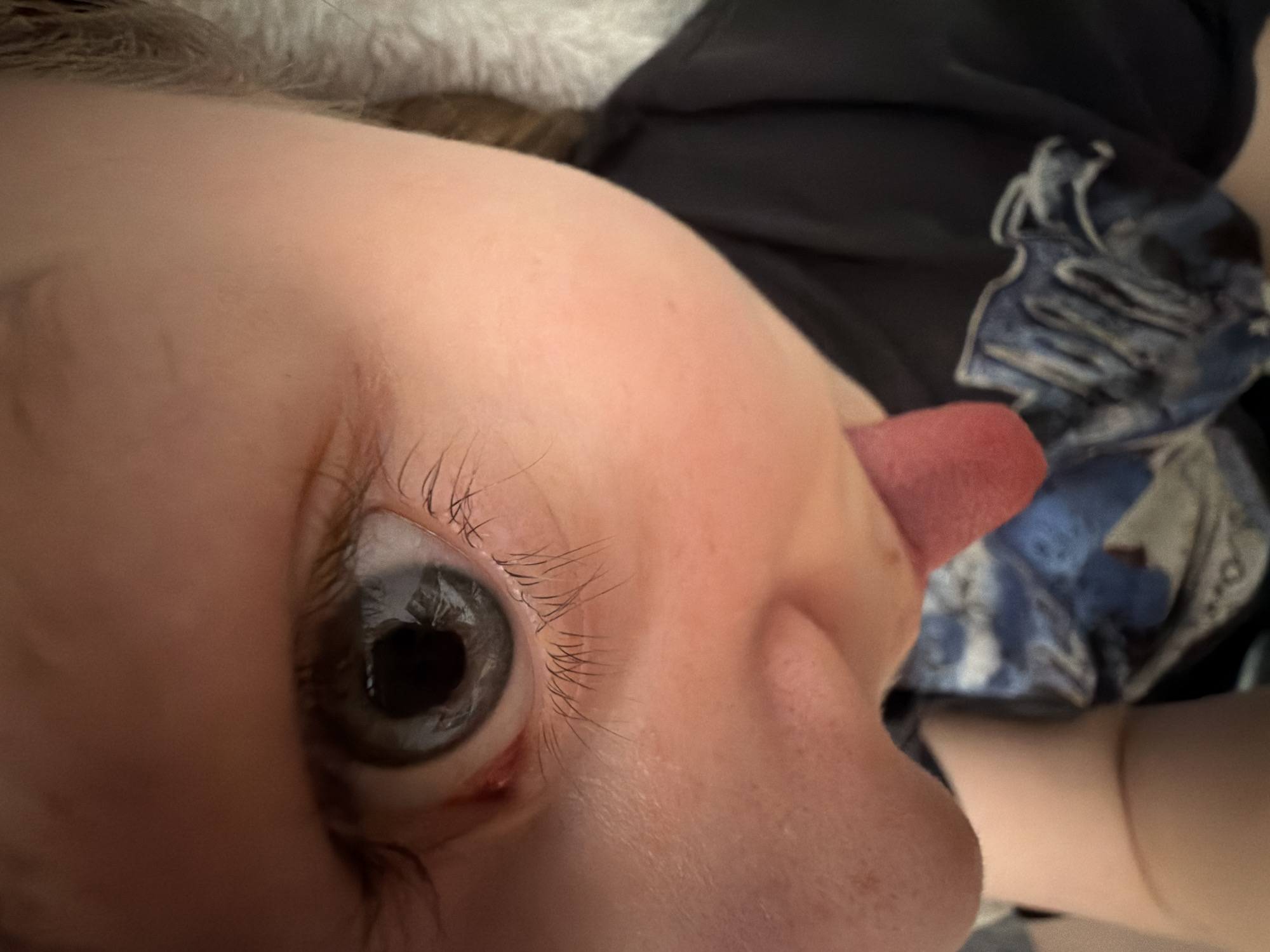 A close-up, low-angle shot captures a segment of a person's face, likely a child's, in what appears to be an indoor setting in Perm, Russia. The dominant feature is a wide-open, light gray-blue eye with a dark, round pupil and long, fine eyelashes. The skin around the eye and a portion of the nose is smooth and light-toned. To the right of the nose, a pink tongue is clearly visible, extended outwards, suggesting a playful or mischievous expression.
The perspective is from below, looking up at the subject, with the camera positioned very close. In the upper left background, a soft, light-colored fabric, possibly a blanket or fur, is partially visible. On the right side of the image, behind the extended tongue, is a dark blue fabric with an abstract, lighter blue and white pattern, likely part of the subject's clothing or a nearby item. The lighting is soft and even, indicating an indoor environment without specific time-of-day cues. There is no visible text in the image. The scene conveys a candid and spontaneous moment of youthful playfulness.