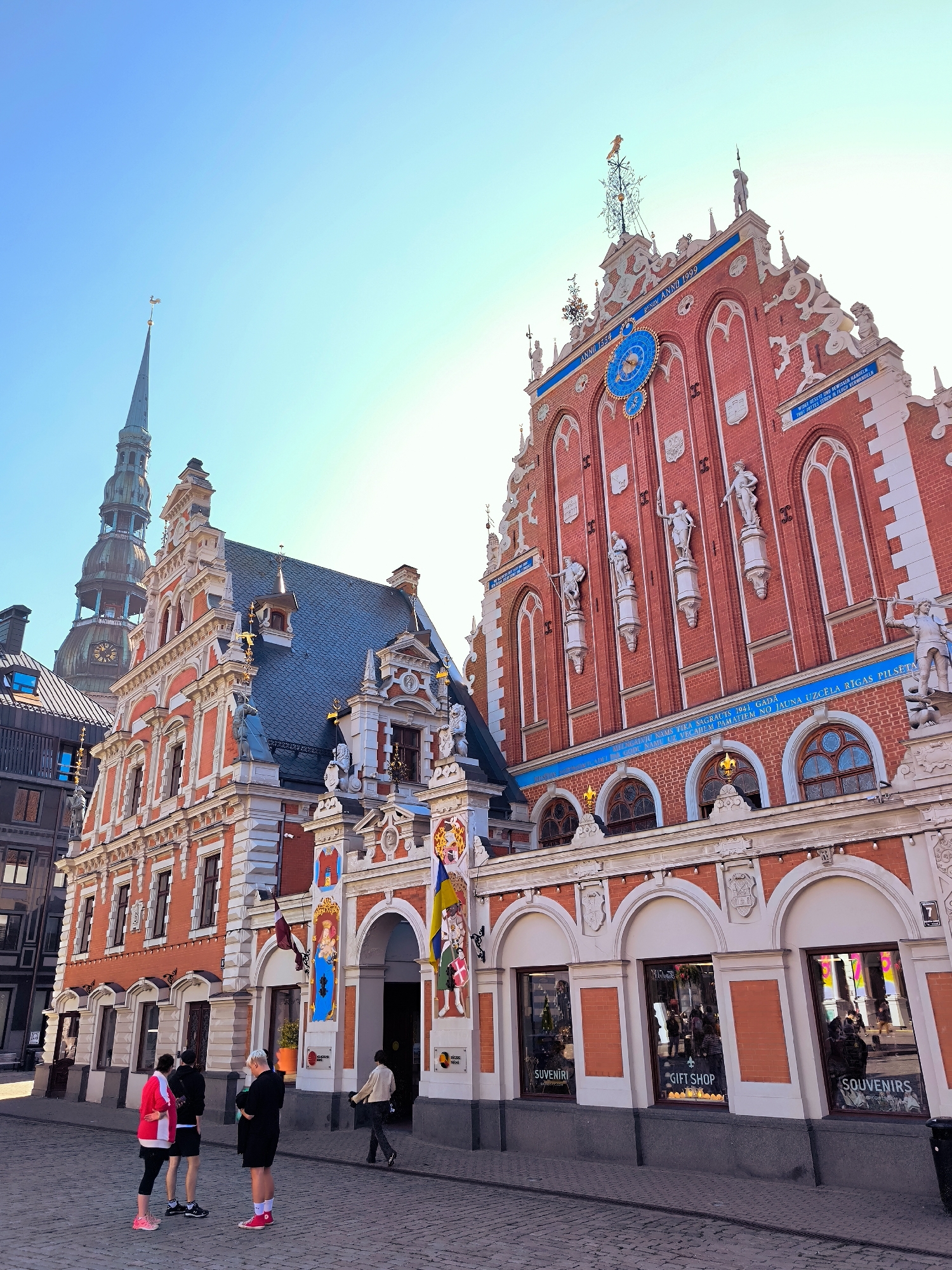 The image shows the House of the Blackheads on Town Hall Square in Riga, Latvia, under a clear blue sky. The historic red brick building, with its ornate white facade and stepped gable, is the primary subject. To the left, another historic building with a tall, slender spire, likely St. Peter's Church, is visible. Several people are present on the cobblestone square. In the foreground, three individuals stand facing away from the camera, looking towards the building. Another person walks further into the square towards an arched entranceway. The ground floor of the House of the Blackheads features shop windows displaying "Souvenirs" and "Gift Shop" signs, with reflections of people and the surroundings visible in the glass. The overall scene depicts a sunny day in a historic European city center.