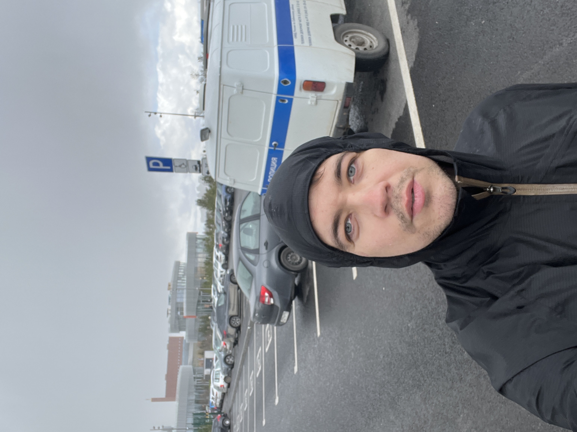 A man in a black hooded jacket takes a selfie in a parking lot in Kazan, Russia, on an overcast day. A white van with "Полиция" (Police) written on it in Cyrillic, with a blue stripe, is parked near the person. There are several parked cars in the lot. A parking sign is visible. Tall buildings can be seen in the background. The ground appears wet.