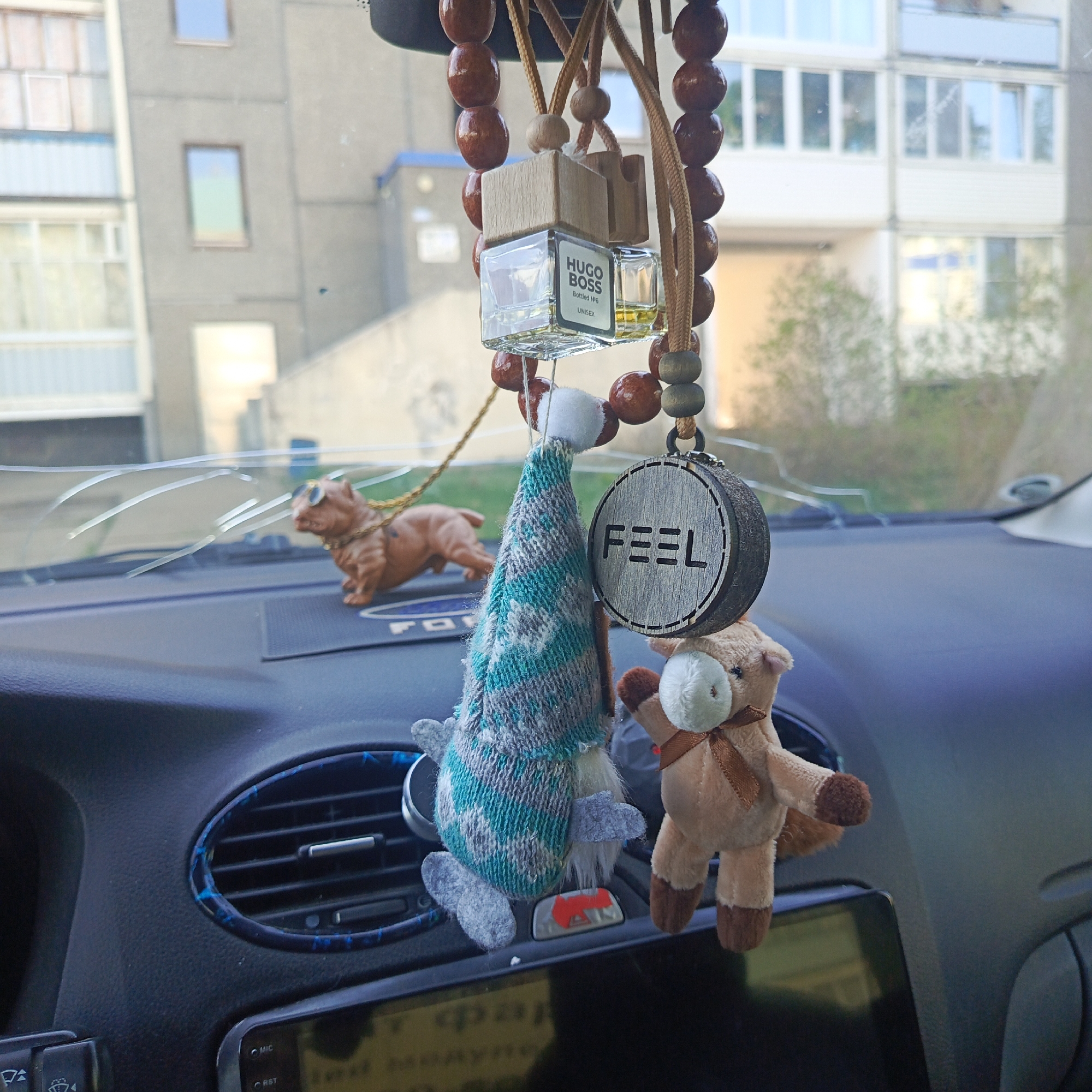 The image is taken from inside a car, looking out through a cracked windshield at a building. The car's dashboard is visible, with a collection of hanging ornaments. These include a miniature Hugo Boss cologne bottle, a wooden charm with the word "FEEL" on it, a gnome-like figure with a patterned hat, and a small stuffed horse. A toy dog wearing sunglasses sits on the dashboard. The cracked windshield obscures the view of the building behind, which appears to be an apartment complex. The location is inferred to be within the city of Марковский, Russia.