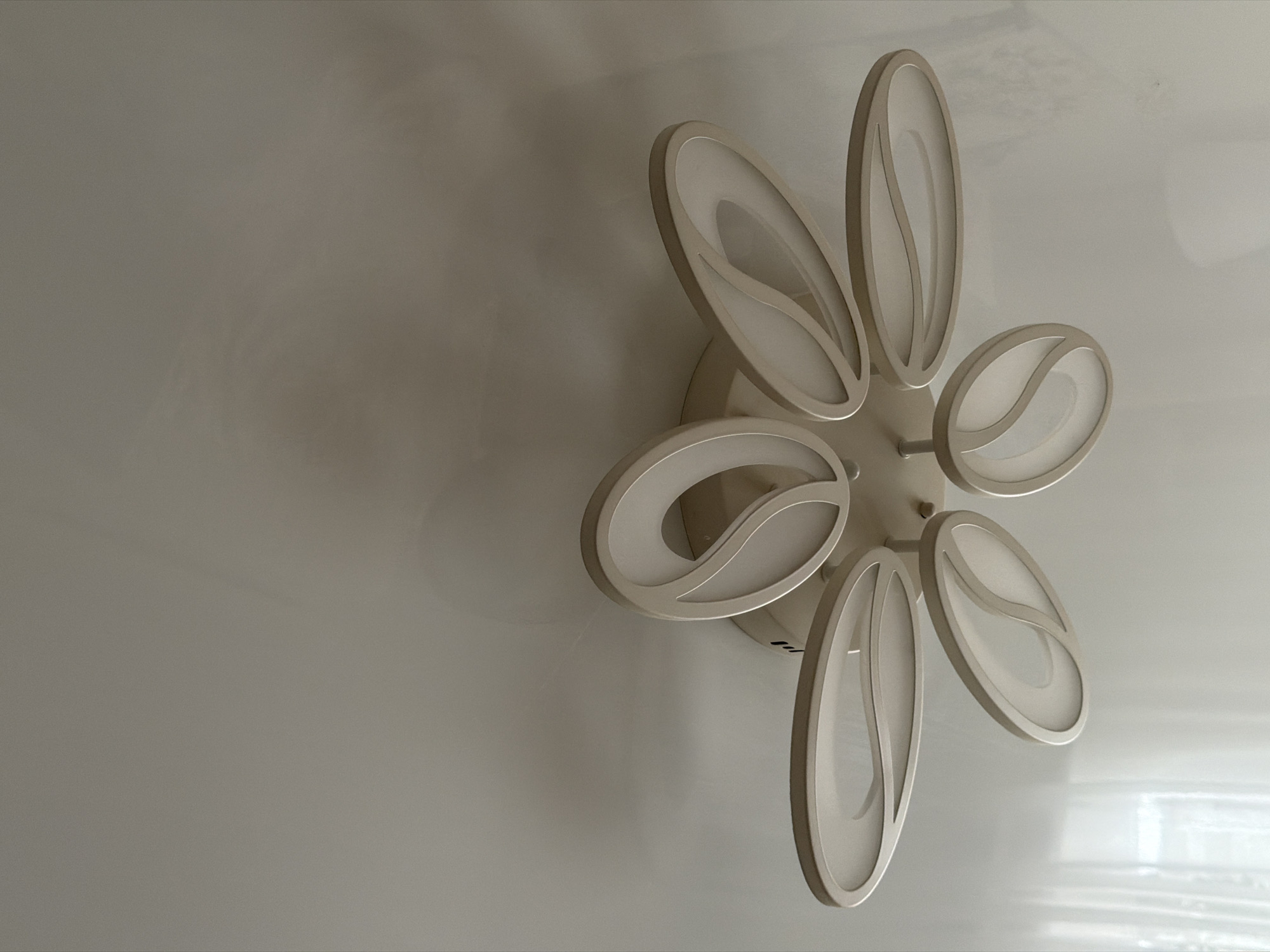 The image shows a modern, white, multi-petaled ceiling light fixture mounted on a white ceiling. The light fixture has a floral or abstract design with several curved, oval-shaped elements that emit a soft, diffused light. The fixture is centrally located in the frame, and the ceiling extends upwards and to the sides. There are no people or other objects visible in the image. The lighting is even, suggesting an indoor environment with ambient light. There is no information about the city of Semiluki, Russia present in the image.