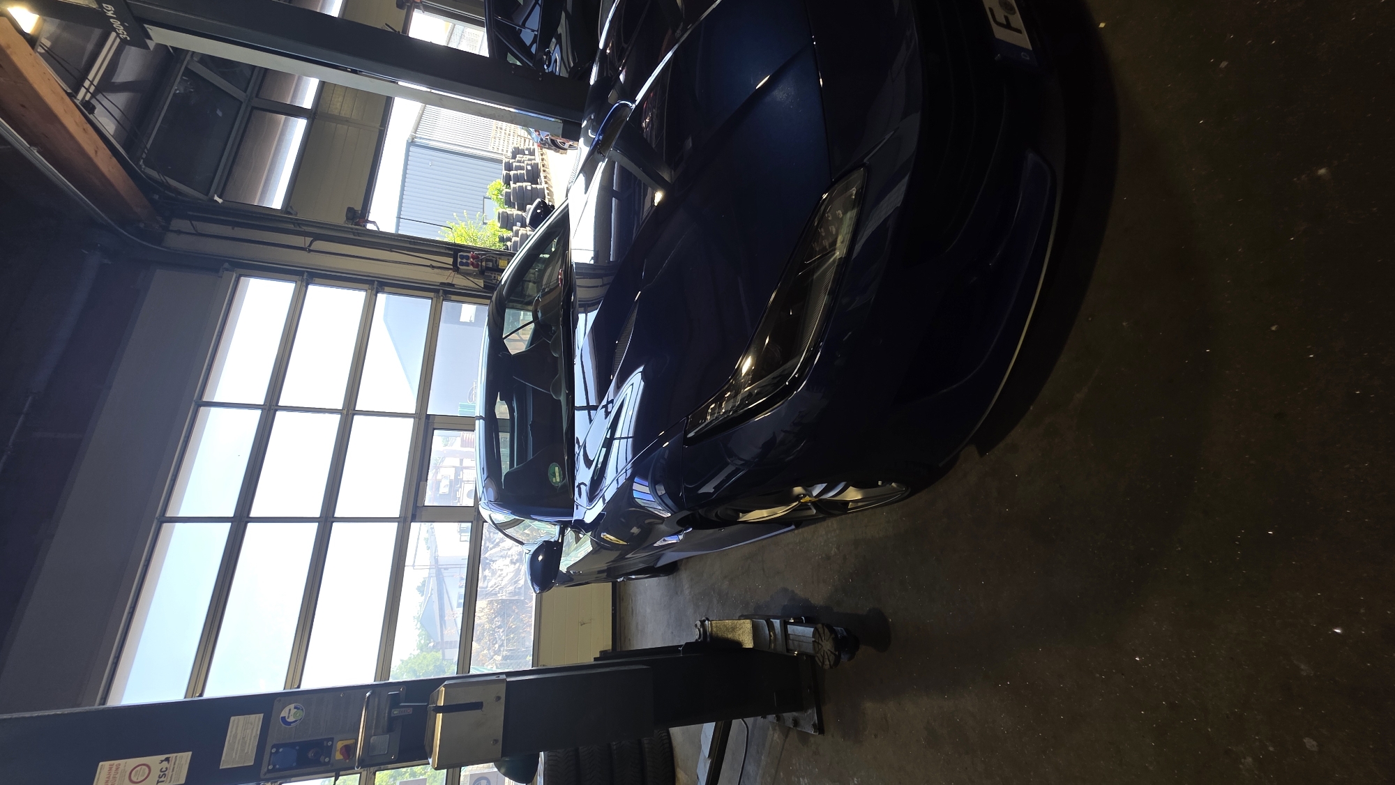 The media file shows a dark blue sports car parked inside a garage or workshop. The car is the main subject, and it is positioned at an angle, with its front end closer to the viewer. The car appears to be sleek and modern, with a low profile and sharp lines. Its headlights are visible, and the bodywork has a glossy finish that reflects the ambient light.
Behind the car, a large window or glass door is visible, allowing natural light to enter the space. The window is segmented into multiple panes, and through it, we can see hints of the outside environment, including what appears to be some greenery and possibly other buildings. The interior of the garage seems to be a concrete floor. A car lift or some other mechanical equipment is also present in the foreground, suggesting that the car is either being serviced or is parked in a facility where such work is done.
The overall lighting suggests it is daytime, possibly a bright day given the strong illumination coming through the windows. There is no visible human activity in the image. The scene evokes a sense of a professional automotive setting.
There is some visible text on an object in the foreground, possibly on the car lift. One visible label reads "3200 KG," indicating a weight capacity. Another label shows a logo with "TSC". There's also a partially visible license plate on the car with the letter "F".