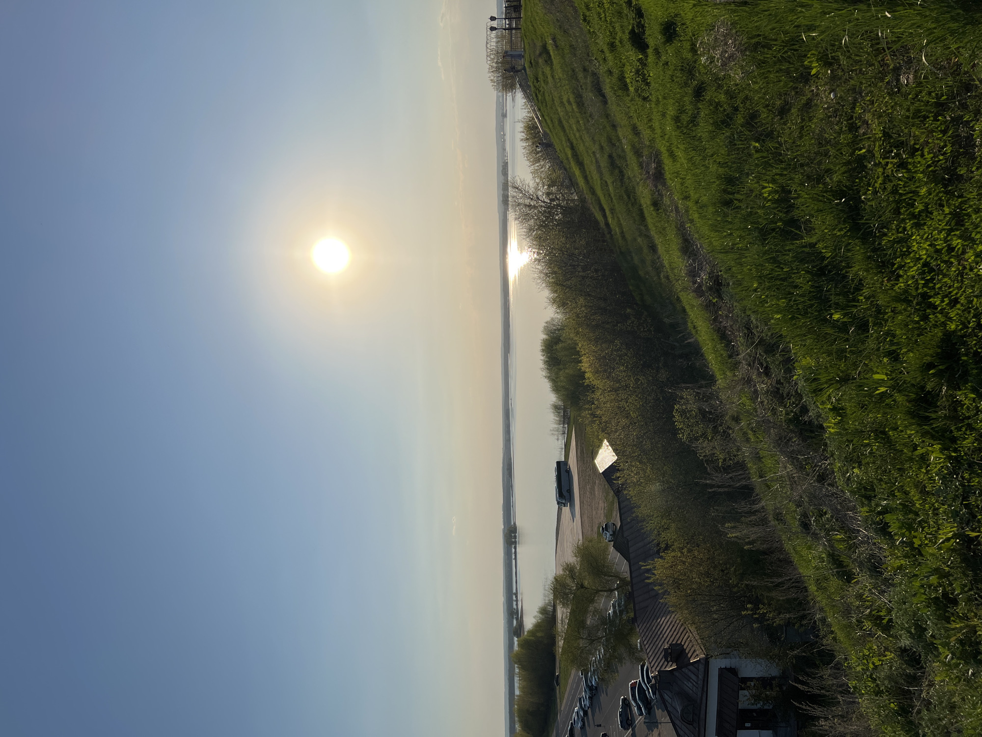 The image shows a view overlooking a landscape in Sviyazhsk, Russia. The sun is high in the sky, casting a glare on the water. A line of trees and vegetation runs along the right side of the frame. A parking lot with cars and a building with a brown roof are visible below the trees. In the distance is a body of water with light reflecting on its surface.