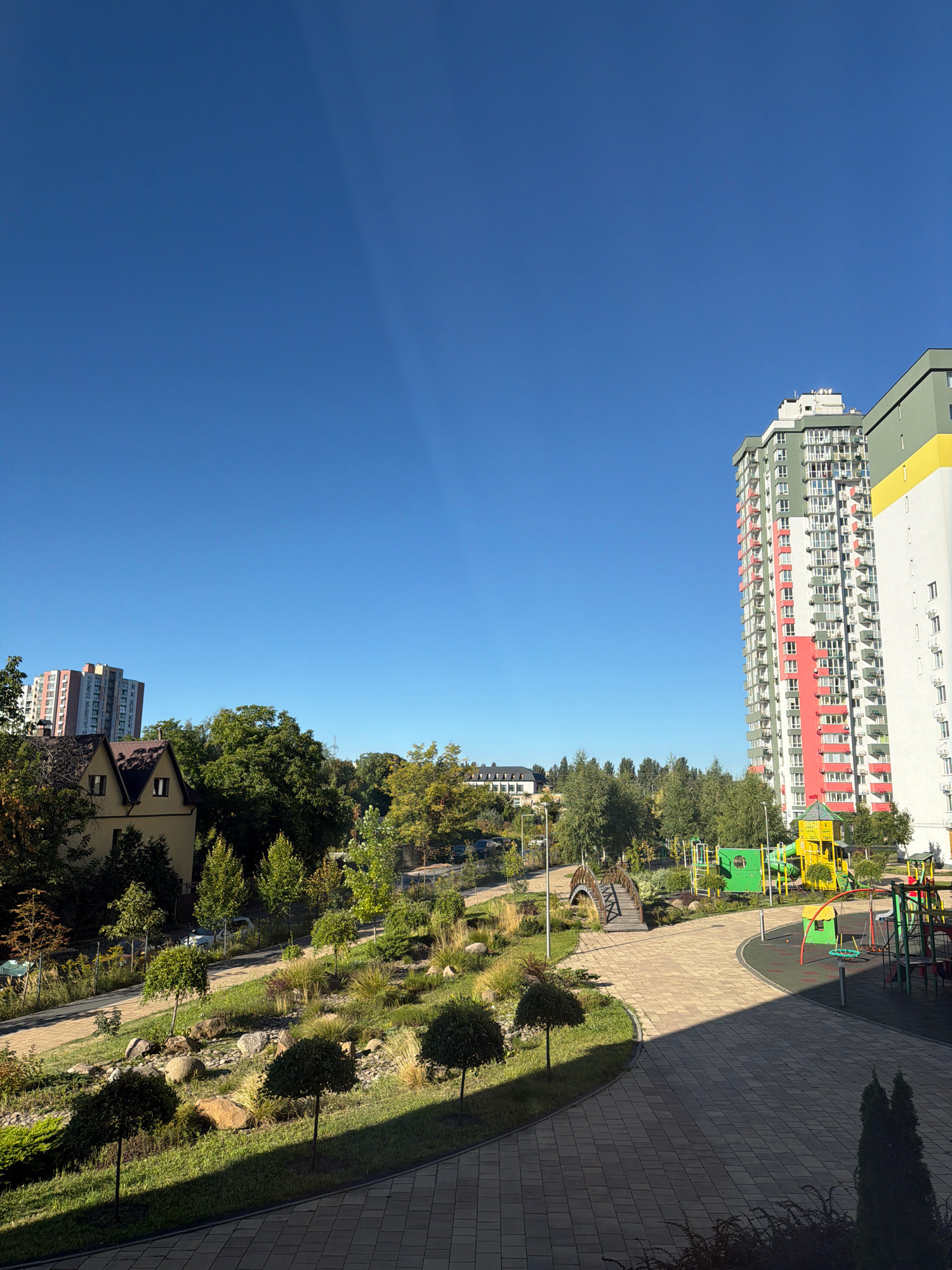 The image captures a bright, sunny day in a residential area of Kyiv, Ukraine. The sky is a clear, vibrant blue, with a faint, soft ray of light stretching across it. In the foreground, a paved walkway with a patterned design transitions into a landscaped area with neatly trimmed trees, rocks, and low-lying vegetation. Sunlight casts distinct shadows across the path and the greenery.
Further into the scene, a playground is visible with colorful structures, including a large yellow and green play set and a red swing set. Beyond the playground, a series of multi-story residential buildings are present. The most prominent is a tall, slender building with a striking facade featuring white, green, and red sections, and numerous balconies. To its right stands another tall building, primarily white with a yellow accent near the top. In the middle ground, nestled among lush green trees, a smaller, traditional-looking house with a brown roof and dormer windows is visible. Another, taller residential building with a lighter facade and pink accents can be seen behind this house. The overall impression is one of a modern, well-maintained urban environment with recreational spaces.