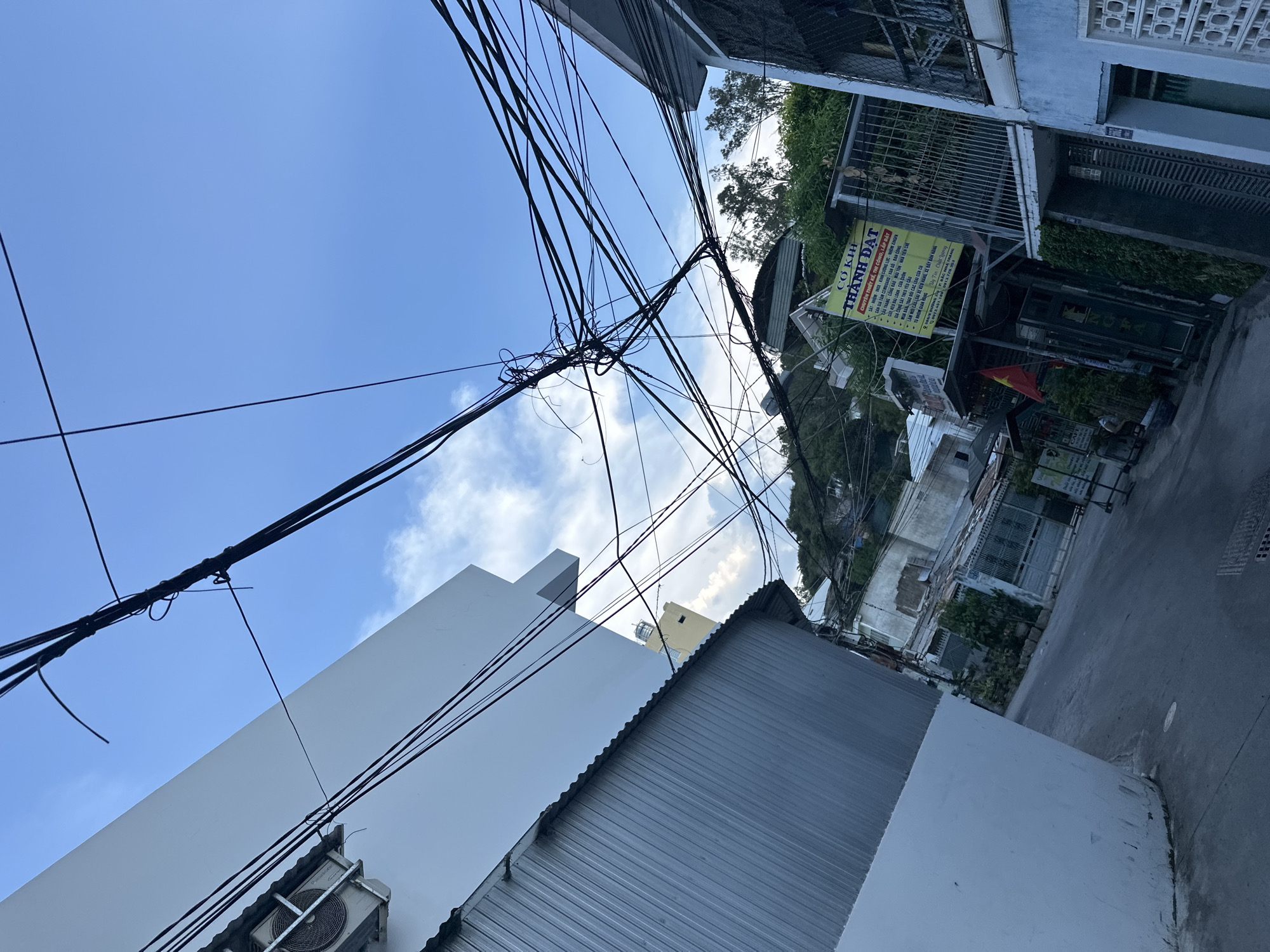 The image captures an urban scene in Bac Nha Trang Ward, Vietnam, characterized by a mix of residential buildings and utility infrastructure. A dense web of overhead power lines dominates the sky. A white building with an air conditioning unit and a corrugated metal roof are visible in the lower left. On the right side, an establishment with a sign that includes "CƠ KHÍ THÀNH ĐẠT" (Mechanical Engineering Thanh Dat) with Vietnamese text beneath, suggesting services related to design, construction, and installation of various metal structures, building components, and related services. The facade includes metal grating, vegetation, and a Vietnamese flag. The street slopes upward with additional buildings and trees visible in the background.