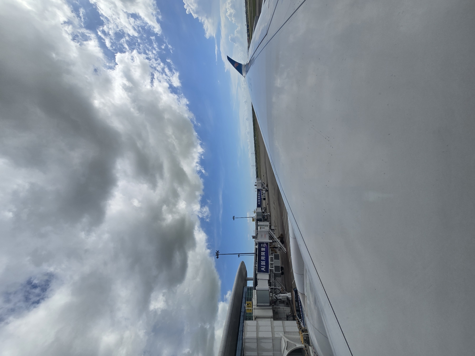 An airplane wing extends into the foreground, reflecting the sky and clouds. The wing is smooth and white, with a visible seam running along its surface. In the background, an airport terminal building is visible, with jet bridges extending out to connect with aircraft. The terminal is a modern structure with a curved roof and large glass windows. The sky is a mix of blue and white clouds, with some patches of darker clouds suggesting potential rain. The scene appears to be a tarmac at an airport, likely Shenzhen Bao'an International Airport in Bao'an District, China, given the presence of Chinese text on the jet bridges. The overall impression is one of an overcast but bright day at an airport.