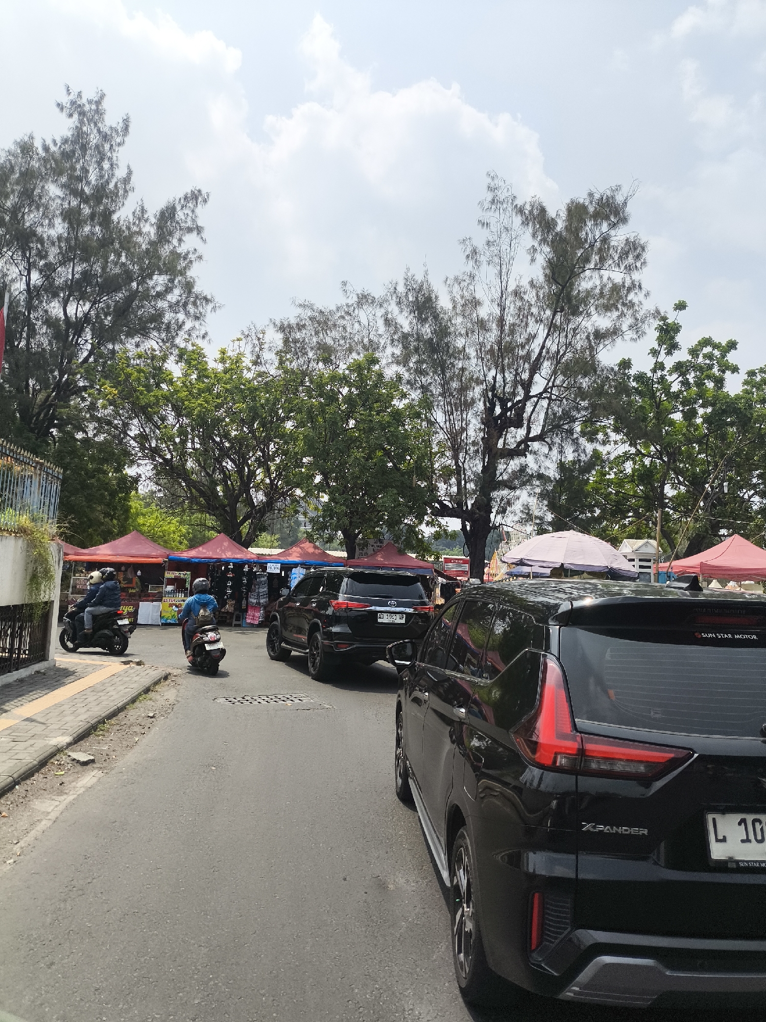 The image captures a street scene in Surakarta, Indonesia, likely during daytime, under a bright sky with scattered clouds. The foreground is dominated by the rear of a black Mitsubishi Xpander SUV, its taillights illuminated. To the left, a street lined with a yellow-striped pedestrian walkway is visible, with a portion of a white building and an ornate metal fence.
Further into the scene, a row of market stalls with red and pink tents are set up along the street, displaying various goods. Several motorcycles are present on the road. One motorcycle, carrying two individuals, is seen on the left side of the frame, moving away from the camera. Another single rider on a scooter is heading towards the market stalls. A black SUV, possibly a Toyota Fortuner, is parked in front of one of the stalls. Lush green trees with tall trunks line the background, creating a canopy over the market area. The overall atmosphere suggests a busy, outdoor market in an urban environment.