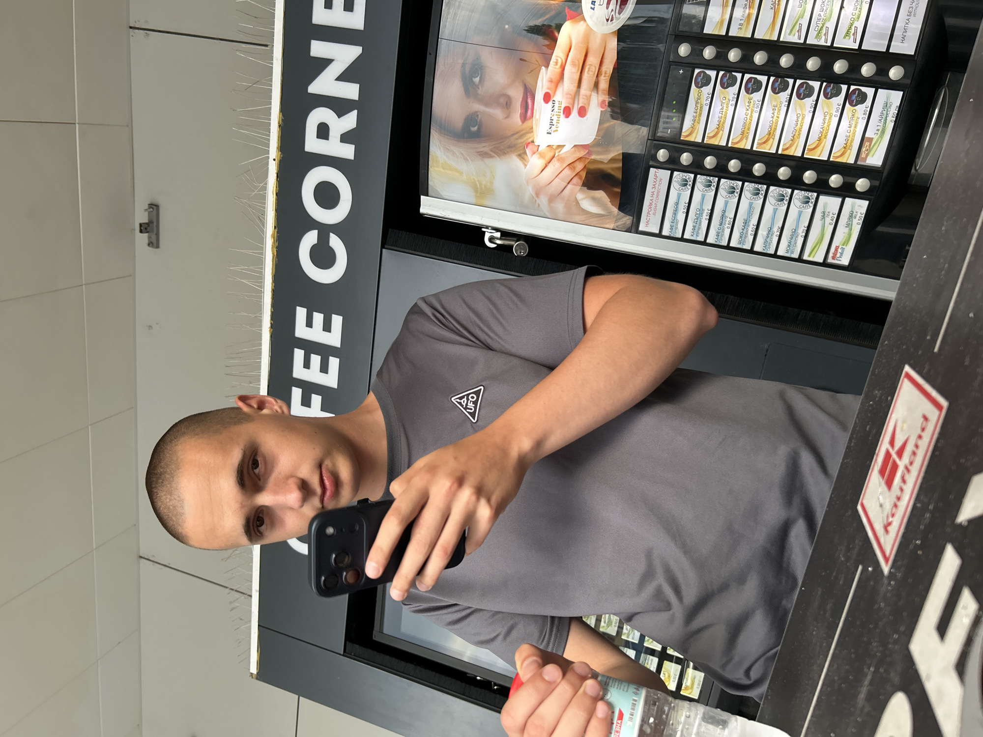 The image shows a young man with a shaved head, wearing a grey t-shirt with a triangular UFO logo on the left chest. He is holding a black smartphone in his right hand and a partially visible plastic bottle of water in his left hand. He is standing in front of a coffee vending machine with various drink options displayed on the screen, including "Espresso Vending." The coffee machine has a promotional image of a woman holding a cup of coffee. Above the machine, a sign reads "COFFEE CORNER." The scene appears to be indoors, possibly in a public space or a store, given the vending machine and the ceiling tiles. There is a "Kaufland" logo visible on a surface in the foreground, suggesting the location might be within a Kaufland supermarket in Vratsa, Bulgaria. The overall atmosphere is casual, with the man seemingly engaged with his phone.