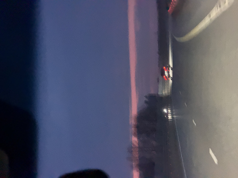 The image depicts a twilight scene in Khanskaya, Russia, observed from the interior of a vehicle looking forward through the windshield. The upper sky is dark blue, transitioning to a distinct horizontal band of pinkish-red light along the horizon. Below this light, a dark, indistinct line of trees is visible on the left, extending towards a flatter, blurry landscape on the right.
The foreground shows a multi-lane asphalt road, appearing damp or reflective, illuminated by the capturing vehicle's headlights. White dashed lane markings are present, along with a continuous yellow line on the right side, indicating a curve in the road.
One dark-colored vehicle, likely a sedan or SUV, is positioned ahead in the right lane, displaying illuminated red taillights and additional white illumination beneath its rear bumper. To the left of this vehicle, a set of bright white headlights from a second, more distant vehicle is discernible. On the far right edge of the frame, multiple blurry, reddish-orange objects, consistent with traffic cones or barriers, are present. No individuals are visible within the frame.