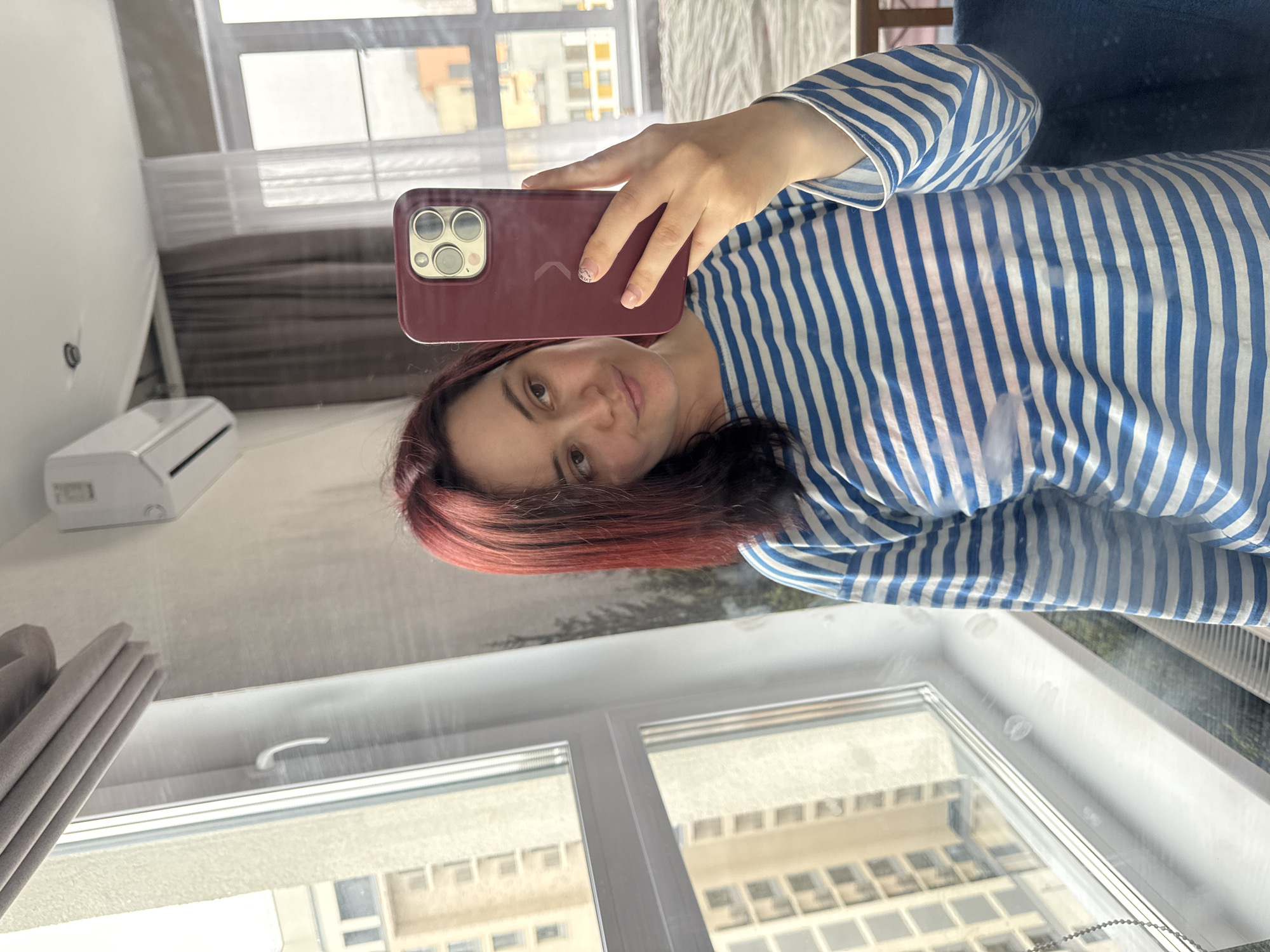 A woman with red-tinted hair is taking a selfie in a room with a window. She is wearing a blue and white striped shirt and holding a burgundy-colored phone. The room has white walls and curtains. An air conditioning unit is visible on the wall. Outside the window, buildings are visible. The scene is located in Izhevsk, Russia.