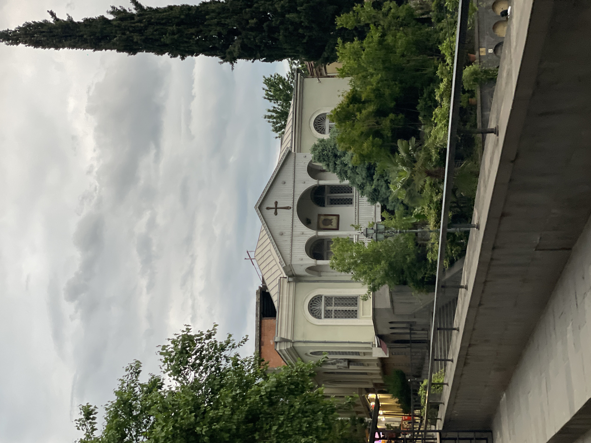 The image captures the Surb Gevorg Church in Tbilisi, Georgia, partially obscured by trees and a bridge. The church, painted a pale hue, features a gabled roof, arched windows, and a cross on its facade. Lush trees surround the structure, while a tall, slender tree stands prominently in the background. The sky overhead is overcast with gray clouds. A concrete bridge with metal railings occupies the foreground, partially obscuring the view of the church. The street below is illuminated with artificial lights. The image is taken from a low angle.