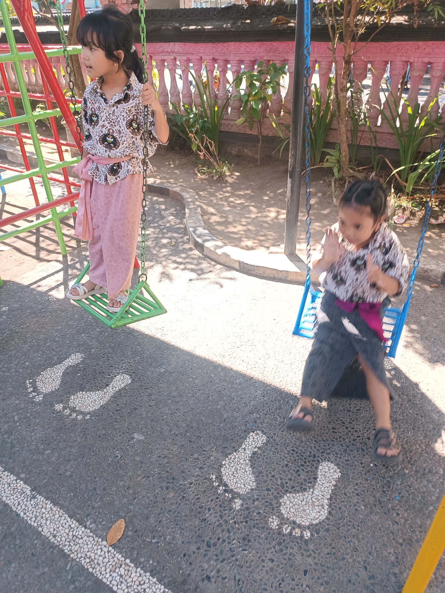 In the city of Tabanan, Indonesia, two young girls are engaged in activity on a playground. The girl on the left, dressed in a patterned shirt and pink wrap skirt, stands on a green swing, holding onto the chains. To her right, a second girl, wearing a similar patterned shirt and a dark wrap skirt, sits on a blue swing, her hands raised in a gesture of play. The playground features a red and green climbing structure on the left and a pink decorative fence in the background, with foliage visible behind it. The ground is paved with dark asphalt interspersed with white pebble footprints, guiding the way. The scene is illuminated by sunlight, casting shadows from the playground equipment and trees.