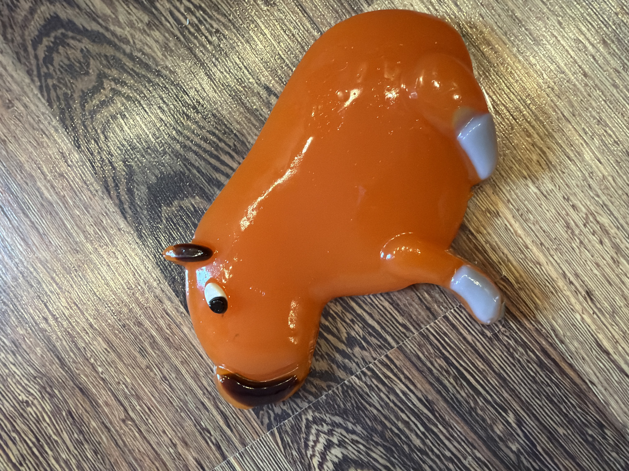 An orange, glossy, capybara-shaped object rests on a wooden floor with a visible wood grain pattern. The object has black eyes with white pupils, small brown ears, and brown accents on its snout. It also has light gray feet. The lighting suggests it's indoors, possibly in a residential setting.