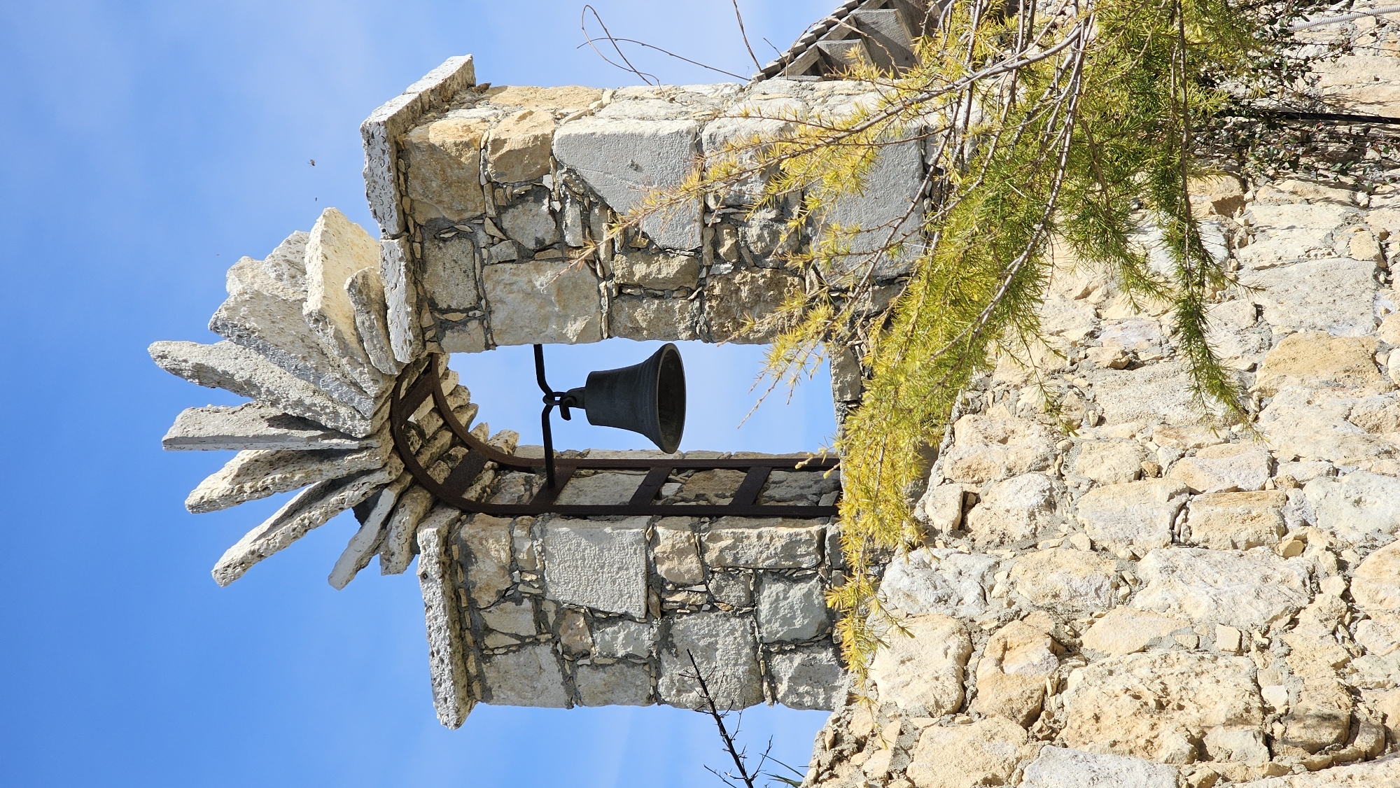 An upright shot captures a historical stone structure, possibly a bell tower, set against a bright, clear blue sky. The main feature is a dark metal bell, centrally suspended by a sturdy metal frame within a rectangular opening of the stone work. The structure is built from light-colored, rough-hewn stones of varying sizes, mortared together.
To the left of the bell's opening, a unique architectural detail is visible: flat, fan-like stone slabs project outwards, creating a textured edge. On the right side of the image, vibrant green and yellow-tinged branches with needle-like leaves hang downwards, partially obscuring the stone wall and sky. A few small, dark specks, likely insects, are visible against the blue sky in the upper left quadrant.
The scene is bathed in bright daylight, indicating clear, sunny weather. There is no human activity depicted, contributing to a tranquil and timeless atmosphere. The stone construction and bell suggest a building with historical significance, fitting the provided context of Varna, Bulgaria. There is no visible text in the image.