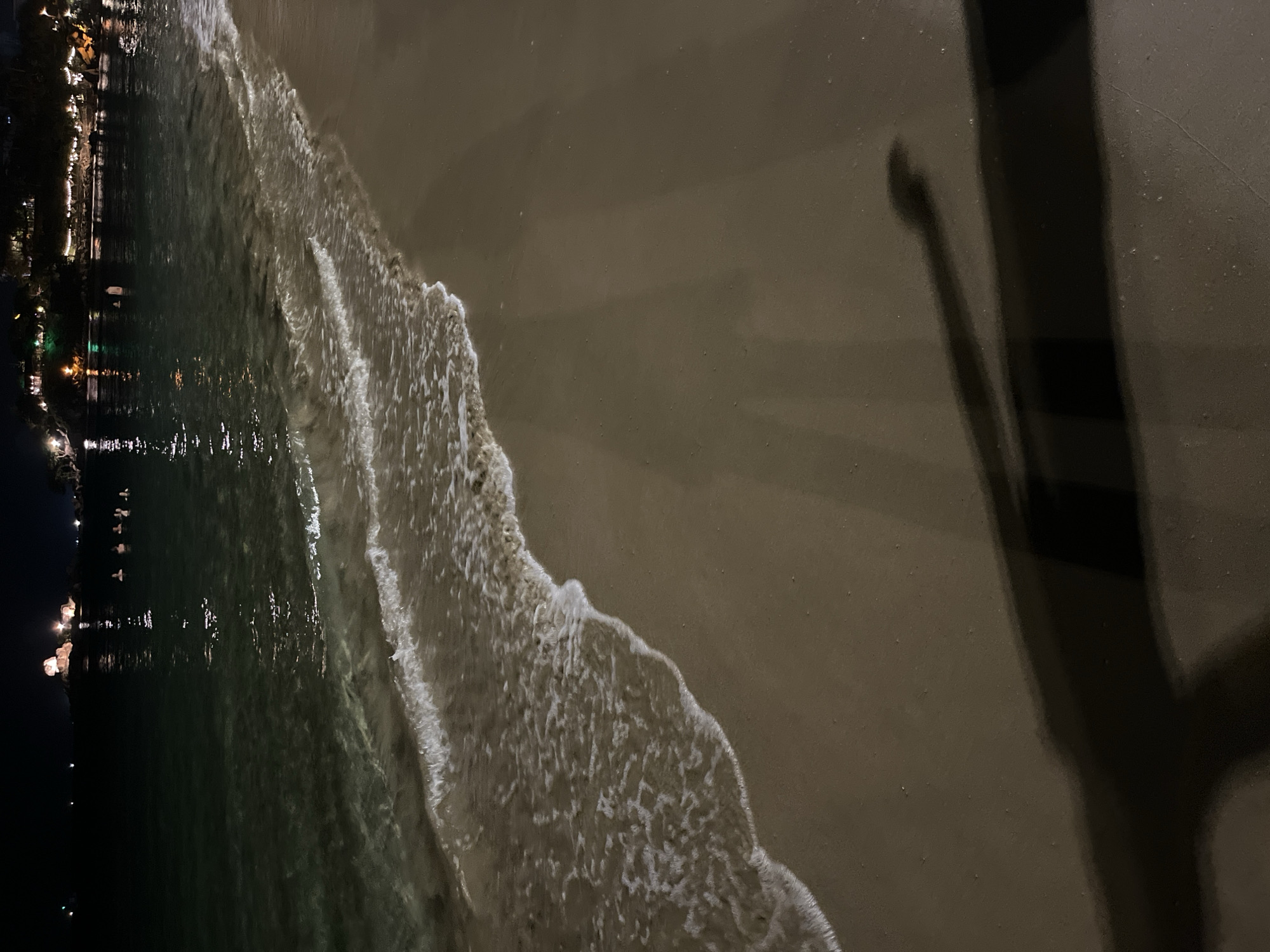 A dark nighttime shot depicts a shoreline. The water is on the left side of the image and is reflecting lights from structures on the shore. A shadow is visible in the upper right of the picture.