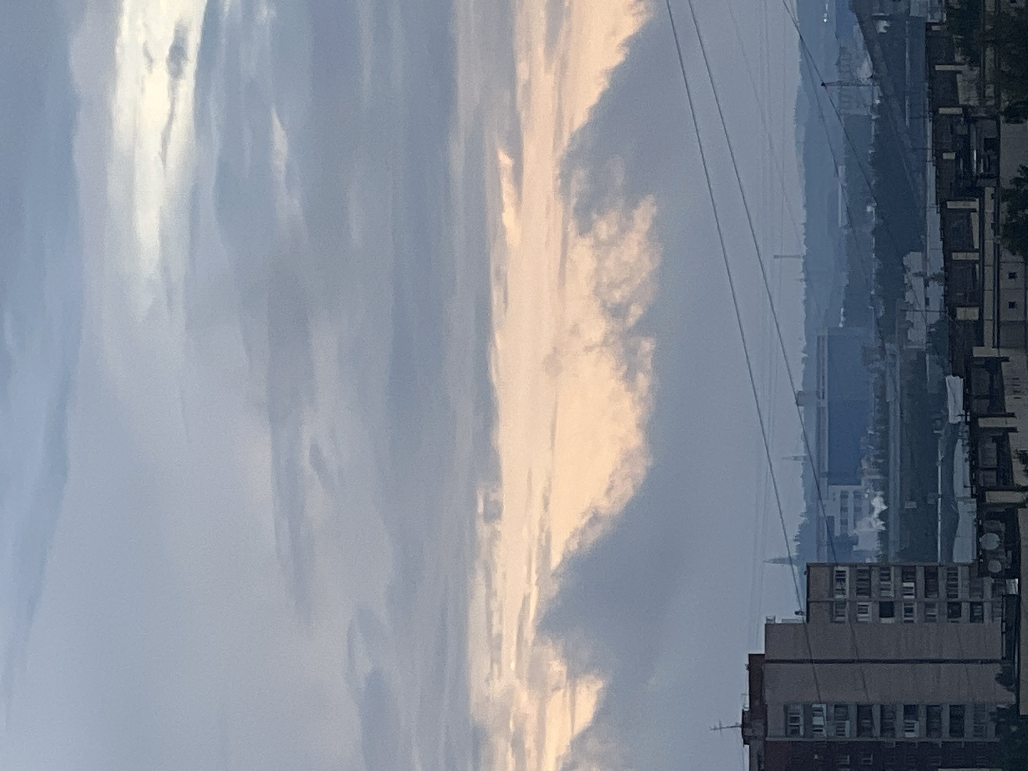 An urban skyline is depicted under a cloudy sky. In the foreground, a tall, multi-story apartment building with visible windows dominates the left side of the frame. Behind it, the cityscape stretches out with various structures, including what appear to be industrial buildings and more residential blocks. Power lines are visible crisscrossing the mid-ground and background. The sky is filled with layered clouds, with a band of warmer, peachy-colored light breaking through in the middle, suggesting either sunrise or sunset. The overall mood is subdued, indicative of overcast weather. The location is identified as Unknown, Russia.