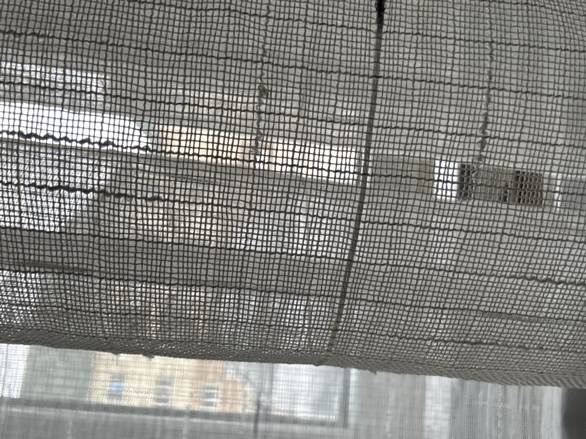 The image captures a view from indoors, looking outwards through a sheer, woven fabric, likely a curtain or blind, which dominates the foreground. This fabric exhibits a prominent grid-like pattern due to its weave, through which light and vague outdoor details are filtered. A thin, vertical cord or string is visible running down the right-center of the image, suggesting it is part of a blind's operating mechanism.
Beyond the textured fabric, a brightly lit outdoor scene is heavily diffused and obscured. The upper part of the background appears almost overexposed due to strong light, indicating daytime, likely a sunny period. In the lower section, through the main fabric and what seems to be another sheer layer, possibly a window screen or a lighter curtain, the indistinct outline of a multi-story building can be perceived. This building features multiple rectangular windows, suggesting an urban or semi-urban environment.
No specific activity or event is taking place; the image presents a static, observational scene. The prevailing bright, diffused light strongly indicates it is daytime. There are no discernible location-specific cues within the image itself to pinpoint Iddo Tudunwada, Nigeria, beyond the general impression of a building viewed from a window. No text is visible in the image. The overall composition highlights the texture of the foreground fabric and the interplay of light and obscurity.