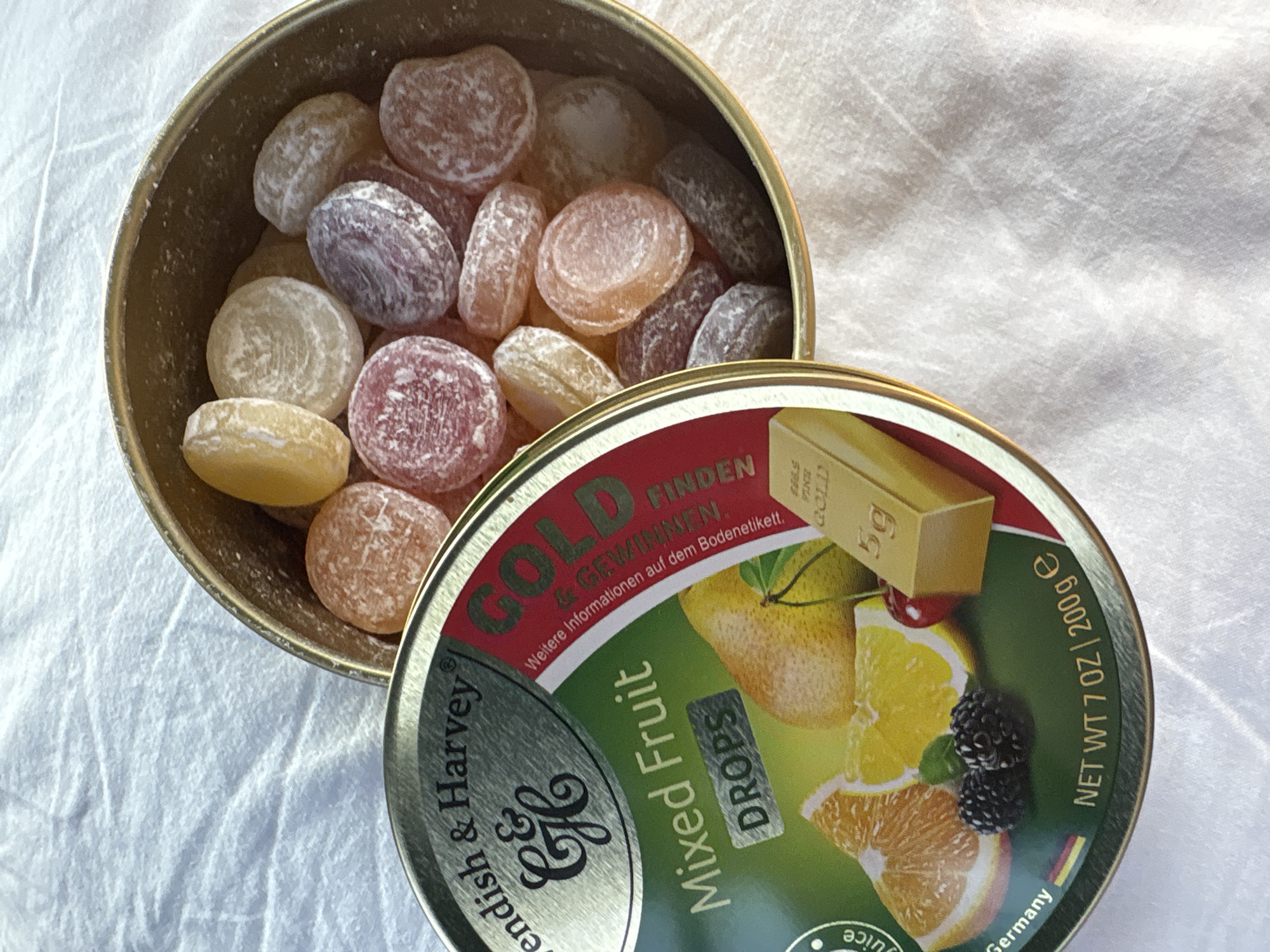 The image shows a round tin of Cavendish & Harvey Mixed Fruit Drops. The tin is open, revealing a pile of colorful, round, hard candies. The lid of the tin, which is partially covering the candies, features an illustration of assorted fruits, including oranges, lemons, and a cherry, as well as two blackberries and a miniature gold bar. Text on the lid indicates "Mixed Fruit DROPS," "NET WT 7 OZ | 200g e," and "Made in Germany." The tin is resting on a white, textured surface, possibly a tablecloth or a sheet. The candies themselves are translucent and appear to be dusted with sugar. The colors of the candies include yellow, orange, pink, purple, and a light brown. The lighting suggests an indoor setting. There are no people or animals visible in the image. The location in Reventin-Vaugris, France is not ascertainable from the image.