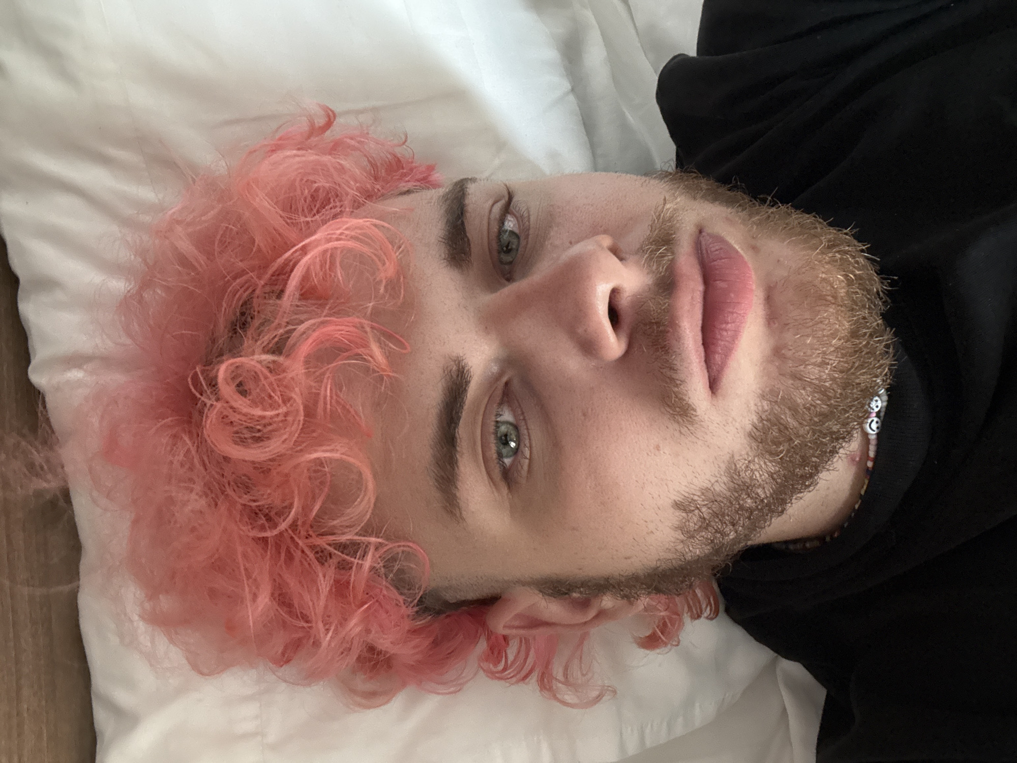 A young man with pink curly hair is lying on a white pillow. He has light skin, green eyes, and a light brown beard. He is wearing a black hoodie. He appears to be indoors. The image does not include any information about the location in Đà Nẵng, Vietnam.