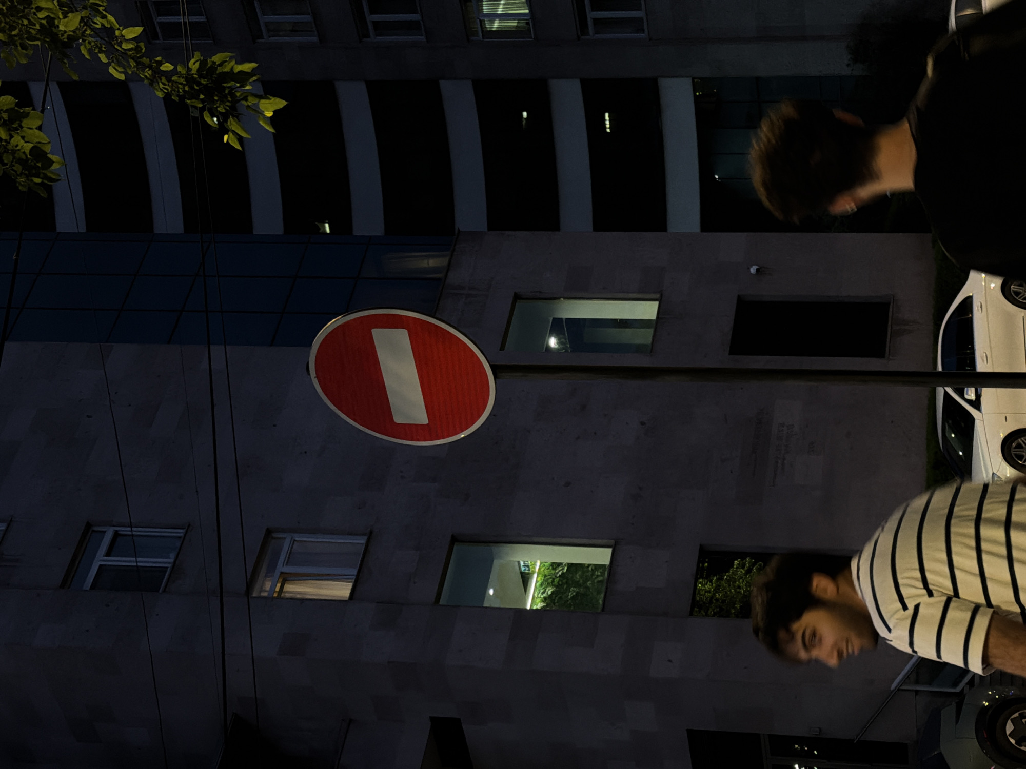 This dimly lit, vertical photograph captures an urban night scene, predominantly featuring a stone or concrete building and two men. The original image appears to be rotated 90 degrees clockwise, so the description assumes a correct upright orientation.
In the center-left, a prominent red circular "No Entry" sign with a horizontal white bar stands out against the dark background of the building. To the far top left, a cluster of green leaves from an unseen tree or bush adds a natural element.
The main background is a multi-story building constructed of dark, textured material, possibly concrete or stone. It features numerous rectangular windows, some dark and reflective, others showing faint interior lights or reflections of foliage. Several larger, dark glass or metal sections are also visible, contrasting with the stone.
Two men are partially visible on the right side of the frame. Towards the top right, a man with short, dark hair is seen from behind, wearing a dark shirt. Towards the bottom right, another man with short, dark hair is shown in profile, wearing a light-colored striped t-shirt. He appears to be looking downwards or to the side. Parts of a white car are visible behind both men, suggesting they are on a street or next to a parking area.
The scene is set at night or late evening, as indicated by the deep shadows, the darkness of the sky, and the artificial lights glowing from some of the building's windows. The activity appears to be casual, with the men simply present in the urban environment. No discernible text other than the universal "No Entry" symbol is visible.