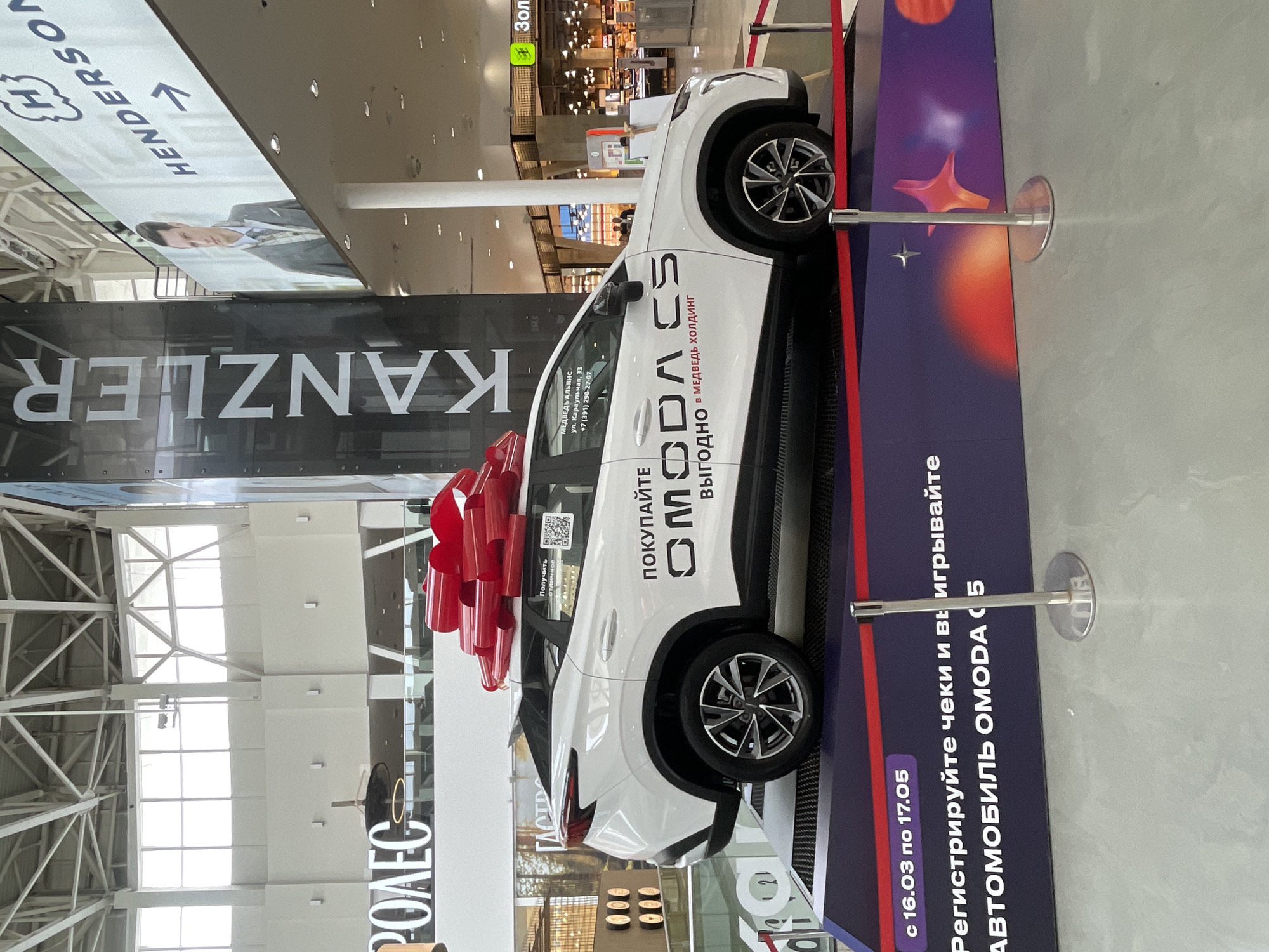 Here is a forensic description of the image:
The image shows a white OMODA C5 car displayed in what appears to be a shopping mall in Krasnoyarsk, Russia. The car has a large red ribbon on it, suggesting it might be a prize or part of a promotion. The car is positioned on a platform with promotional material: "BUY OMODA C5 PROFITABLY IN MEDVED HOLDING" and "Register the receipts and WIN THE OMODA C5 CAR from March 16th to May 17th".
Around the car, there are rope barriers held up by stanchions to keep onlookers at a distance. The background of the shot shows other mall elements like store signs (HENDERSON, KANZLER) and mall interior structures. The mall appears spacious and brightly lit. There are no people immediately visible in the foreground, but the blurred background suggests the presence of shoppers in the mall.