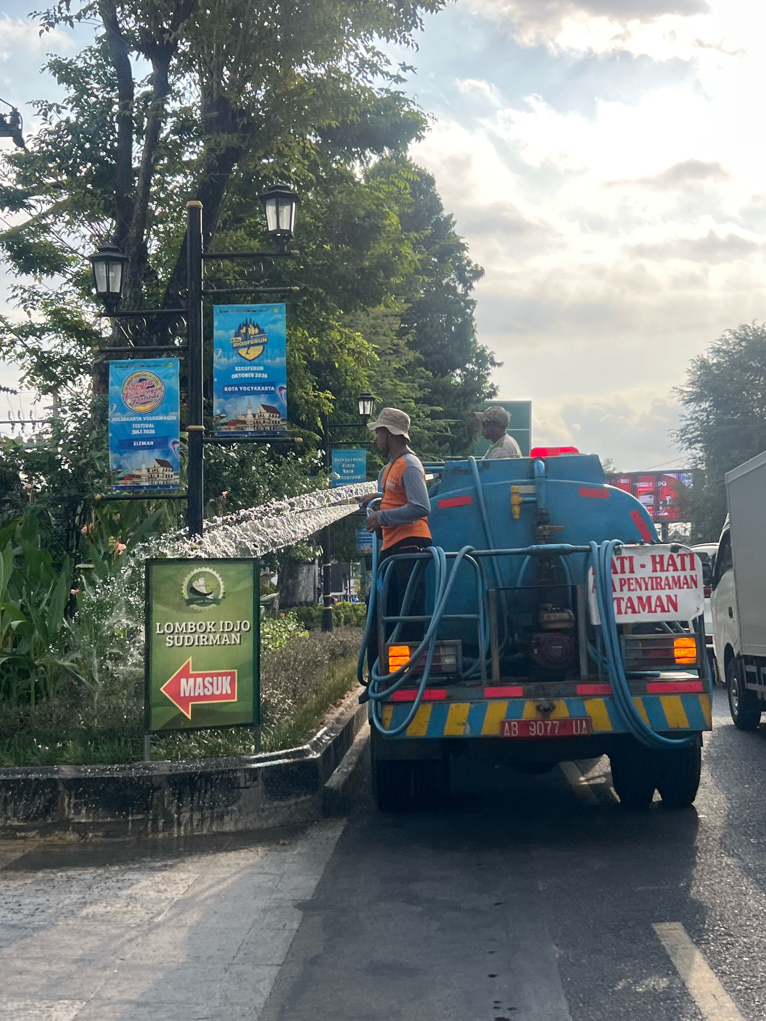 Two men are working on a blue water truck on a street in Yogyakarta, Indonesia. One man, wearing an orange vest and a beige hat, stands on the back of the truck and sprays water from a hose towards the left side of the image. The second man, wearing a lighter colored shirt and a cap, is also on the back of the truck, observing. The truck has a license plate that reads "AB 9077 UA". Next to the truck, a green sign with a red arrow points to the left and displays the text "LOMBOK IDJO SUDIRMAN" and "MASUK". In the background, there are trees, streetlights with banners, and other vehicles. One banner on a streetlight advertises "BIOSFERUN OKTOBER 2025 KOTA YOGYAKARTA". Another banner shows "YOGYAKARTA VOLKSWAGEN FESTIVAL". The sky is partly cloudy, with the sun visible. The scene appears to be a street in a city with greenery, suggesting a well-maintained urban environment.