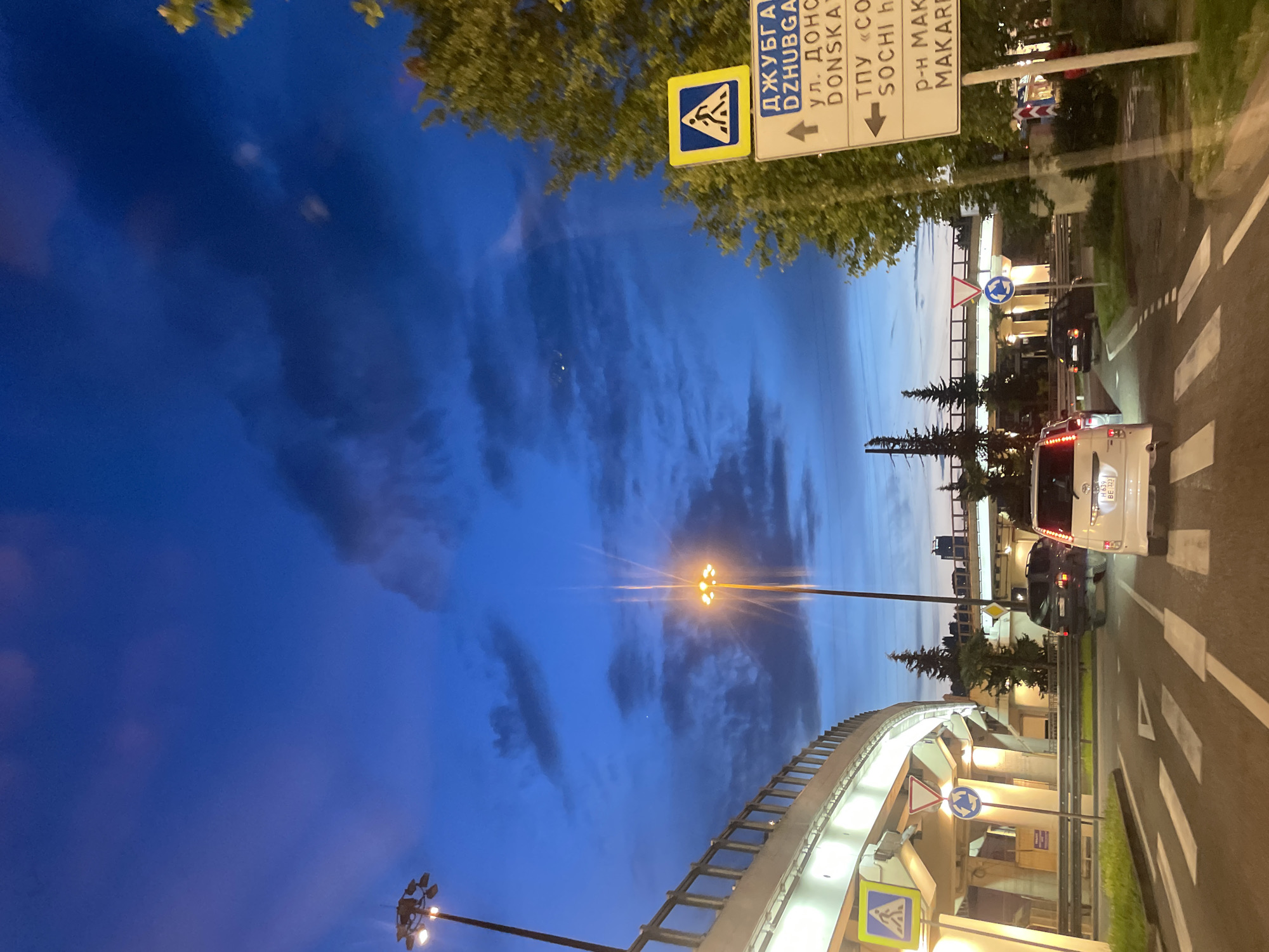 The image depicts a street scene in Sochi, Russia, at dusk. The street is lined with trees and streetlights, with a raised walkway and a road with cars traveling on it. Street signs indicate the directions for "Dzhubga", "Donska Street", "Sochi", and "Makarenko District." The sky is a deep blue with scattered clouds. A crosswalk is painted on the road, and cars are stopped at an intersection. The overall impression is that of a typical evening in a Russian coastal city.