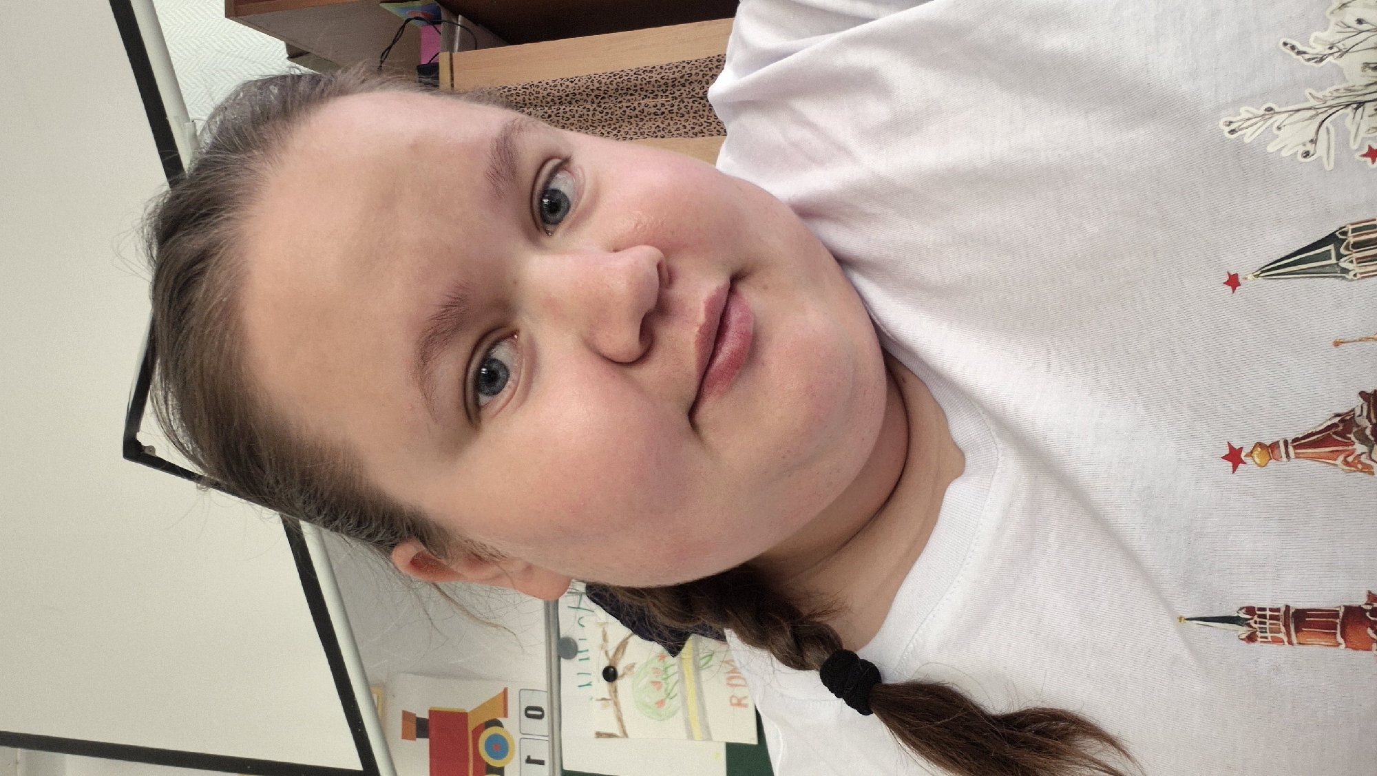 A close-up, slightly angled portrait of a young woman with fair skin and light blue eyes. Her brown hair is pulled back, with a braid visible on the right side. She is wearing a white t-shirt with an image of the Kremlin Towers on it. The background is blurred, but includes glimpses of a room with a whiteboard, furniture, and what appears to be children's artwork. There is no indication in the image that could place this image in the city of Chaikovsky, Russia.