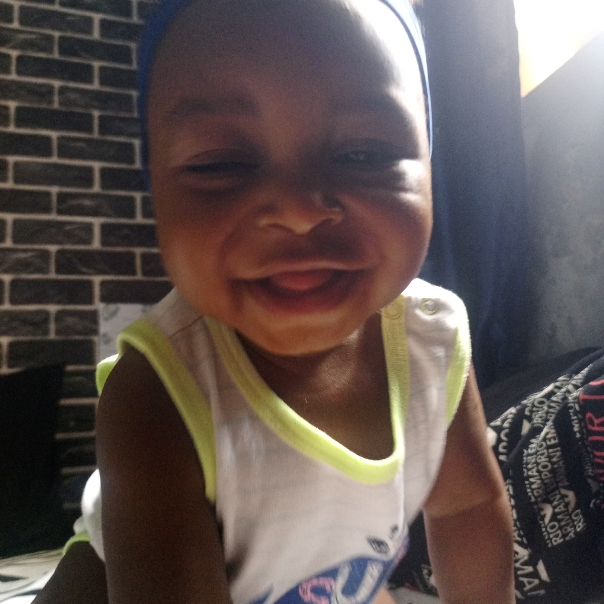 A close-up indoor portrait captures a happy baby or toddler, likely in Keffi, Nigeria. The child, with dark skin and a blue headscarf or headband, is positioned centrally, looking directly at the camera. A wide, joyful smile reveals the tip of their tongue. One eye is slightly squinted, possibly due to bright light or the expression.
The baby is wearing a white sleeveless top with a prominent neon yellow trim around the neck and armholes. The top also features a faint graphic design, partially visible, in shades of blue and pink near the bottom.
The background is split. To the left, a dark brick-patterned wall is visible, suggesting an interior setting. To the right, a dark blue or grey curtain or wall is present, with a bright, indistinct light source casting a glow from the upper right, hinting at daytime. In the lower right corner, a dark fabric is visible, patterned with repeating white text that includes the words "ARMANI EM... EMPORIO ARMANI EM... ARMANI". The overall impression is one of warmth and happiness.
