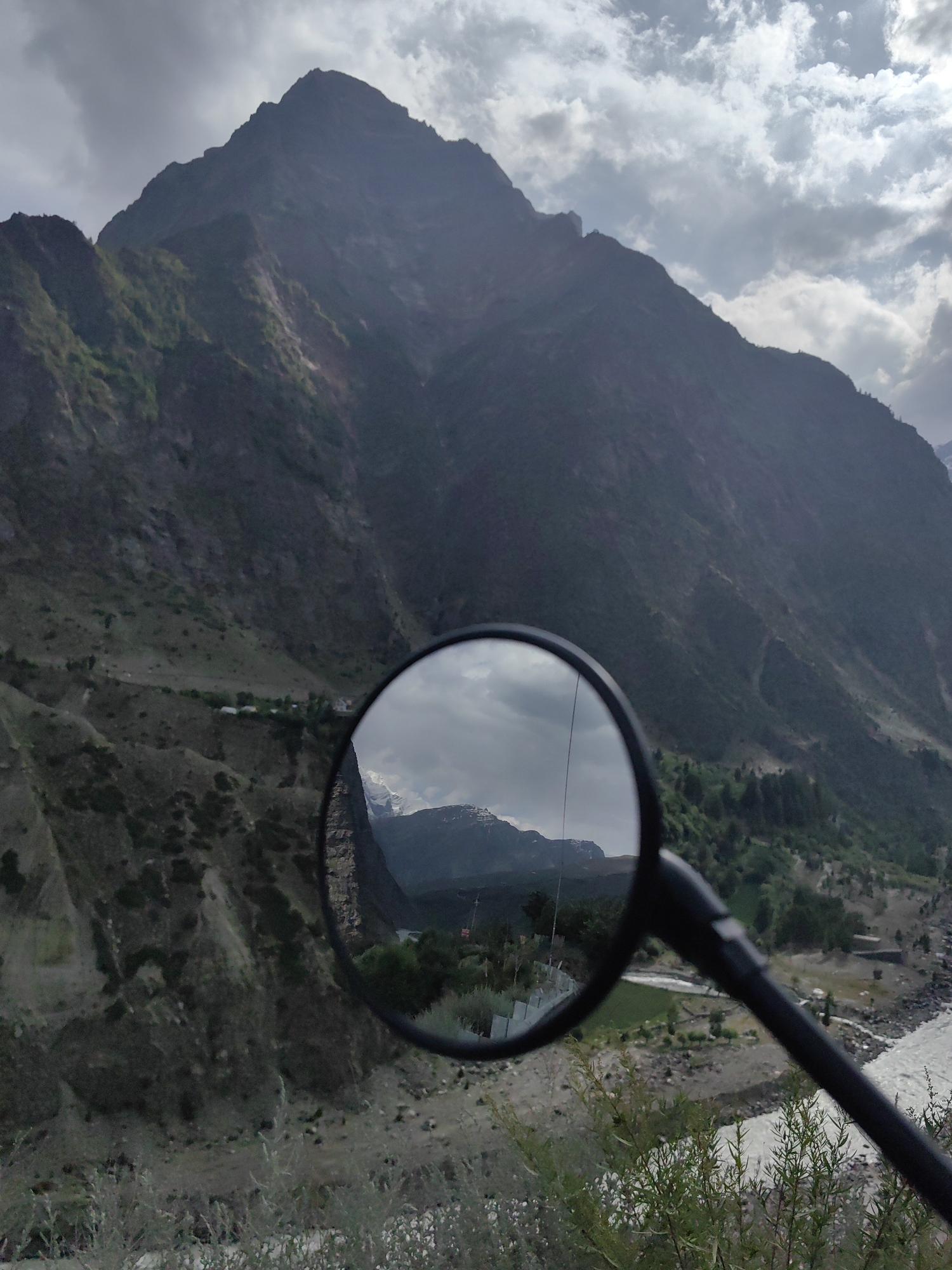 The image captures a mountainous landscape, likely in the vicinity of Kirting, India, as viewed through a motorcycle's rearview mirror. The primary subject is a large, dark mountain with rugged slopes and patches of green vegetation, dominating the upper half of the frame. The sky is overcast with dramatic clouds, suggesting a moody or potentially stormy atmosphere.
The rearview mirror, positioned in the lower half of the image, reflects a section of the landscape. In the reflection, a distant mountain range with snow-capped peaks is visible against a cloudy sky. Below these peaks, the reflection shows a valley with scattered buildings, green fields, and a river. The river appears wide and fast-flowing, with turbulent water.
The foreground of the image, outside the mirror's reflection, shows the textured terrain of a mountainside with dry vegetation and rocks. The lower right corner also shows the edge of the motorcycle's handlebar and a glimpse of more green foliage.
The overall scene conveys a sense of remote, natural beauty and the experience of traveling through a mountainous region. There are no people or animals visible in the image. The lighting is subdued due to the overcast conditions.