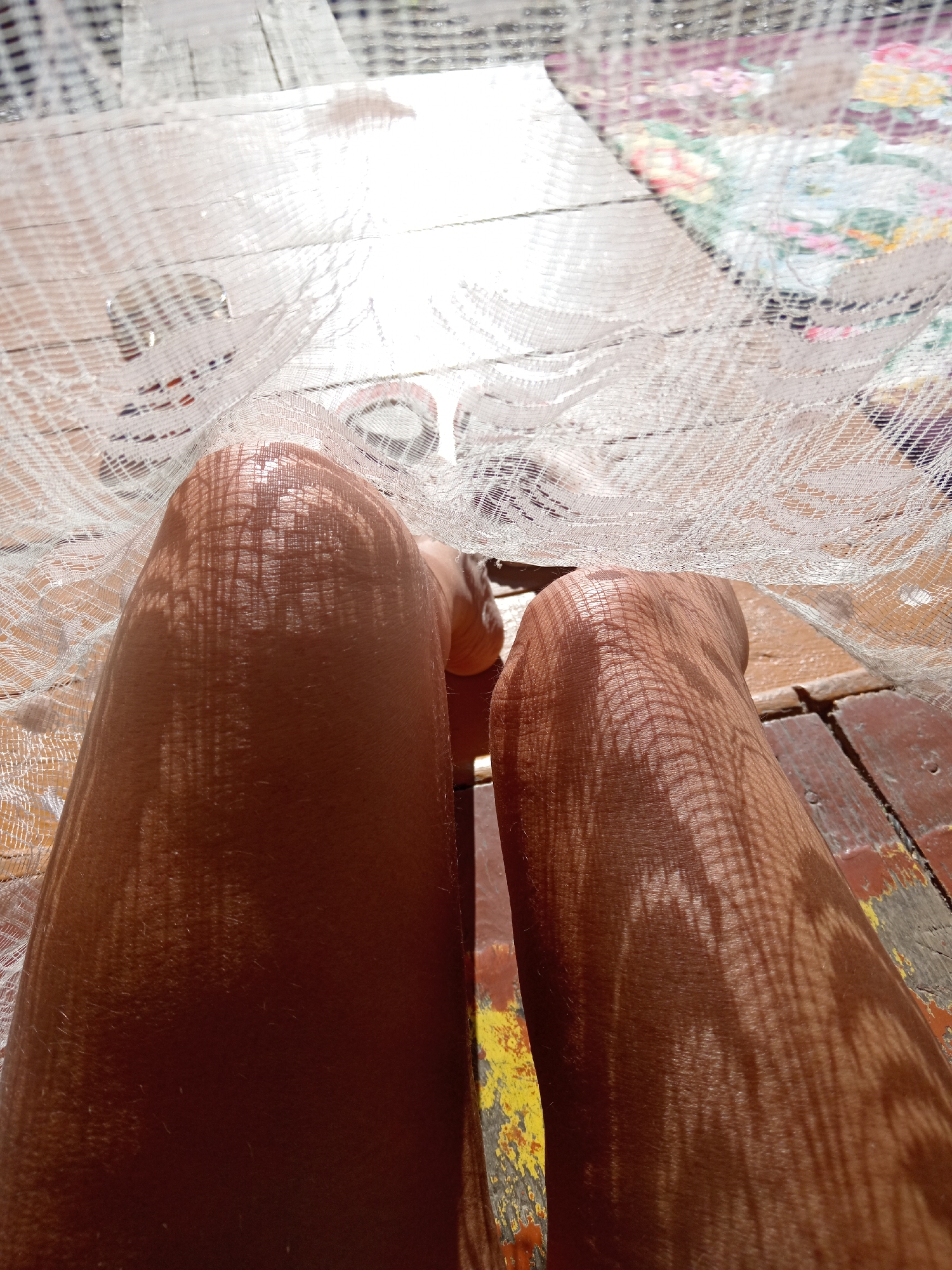 The image shows a close-up view of a person's legs, seen from a low angle looking upwards. The legs are tanned and appear to be bare. They are positioned on a wooden surface that looks like a deck or porch, with planks of varying colors, some appearing worn and chipped. Over the legs, a white, lacy fabric is draped, casting a patterned shadow of crisscrossed lines onto the skin. The fabric is somewhat translucent, allowing light to pass through and creating bright highlights. In the background, beyond the fabric, hints of a patterned surface, possibly a rug or tablecloth, can be seen. The scene suggests a sunny day, likely outdoors, where the lacy fabric is acting as a shade or decorative element. The location is described as the city of Novopokrovka, Russia.