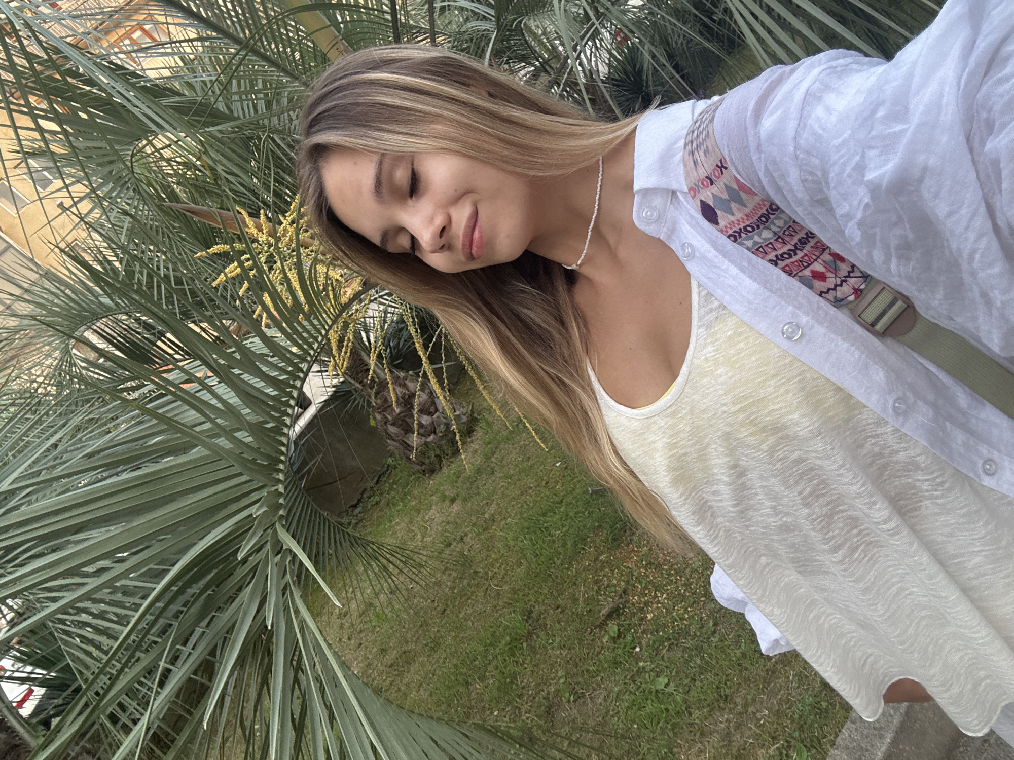 An outdoor, medium-shot, eye-level, selfie taken by a young Caucasian woman with long, straight, blonde hair and closed eyes, wearing a white button-up shirt over a yellow tank top, and a delicate beaded necklace. She is positioned in front of large, green palm fronds with a grassy area and building in the background, suggesting a location in the city of Sokhumi, Abkhazia. The sunlight is diffused, casting a soft glow on the scene. The woman's head is tilted slightly upwards, and her face has a serene expression. A patterned strap is visible crossing her chest over the white shirt. The composition is tightly framed, focusing on the woman and the surrounding greenery.