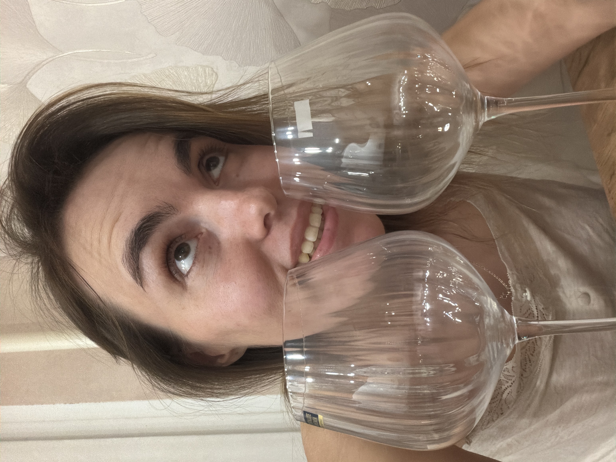 A woman is holding two empty wine glasses in front of her face. She is smiling, and her face is partially obscured by the glasses. The woman has long brown hair and is wearing a light-colored top. The background is a light-colored wall with a subtle pattern. The location is not discernible from the image, and there is no indication of it being in Izhevsk, Russia.