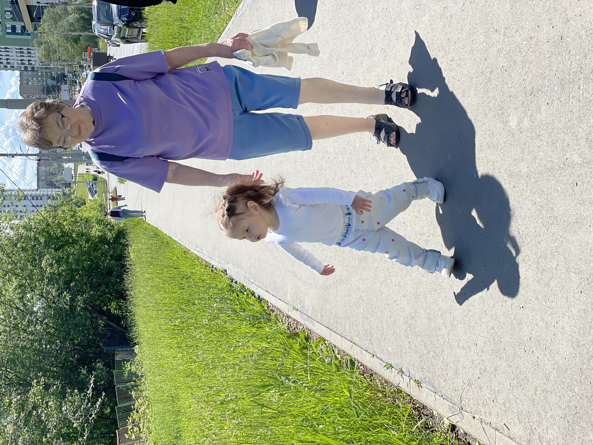 The photograph shows an adult and a young child standing on a paved walkway in what appears to be an urban setting. The adult, positioned above the child in the frame, is wearing a purple t-shirt and blue shorts, with dark sandals. They hold a white knitted garment in one hand. The child, dressed in a white long-sleeved shirt and patterned pants, is positioned in front of the adult. Shadows cast on the walkway suggest a sunny day. Green grass and foliage are visible along the edge of the walkway. In the background, buildings are visible, indicating a city setting. According to information gleaned from the surrounding area, this location is in Izhevsk, Russia.