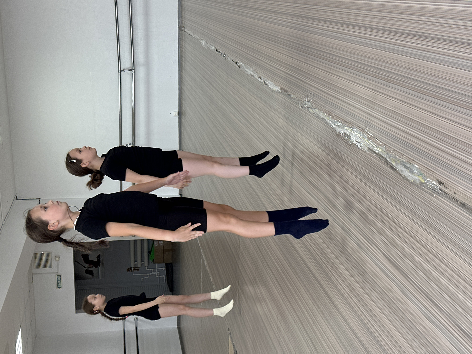 The image shows three young girls in a mirrored room. They are wearing black shorts and tops, with different coloured socks. The girls are lying on the floor facing upwards in a dance studio in Votkinsk, Russia. A barre is visible on the wall behind them. The room has a large mirror running along one side and a white wall on the other.